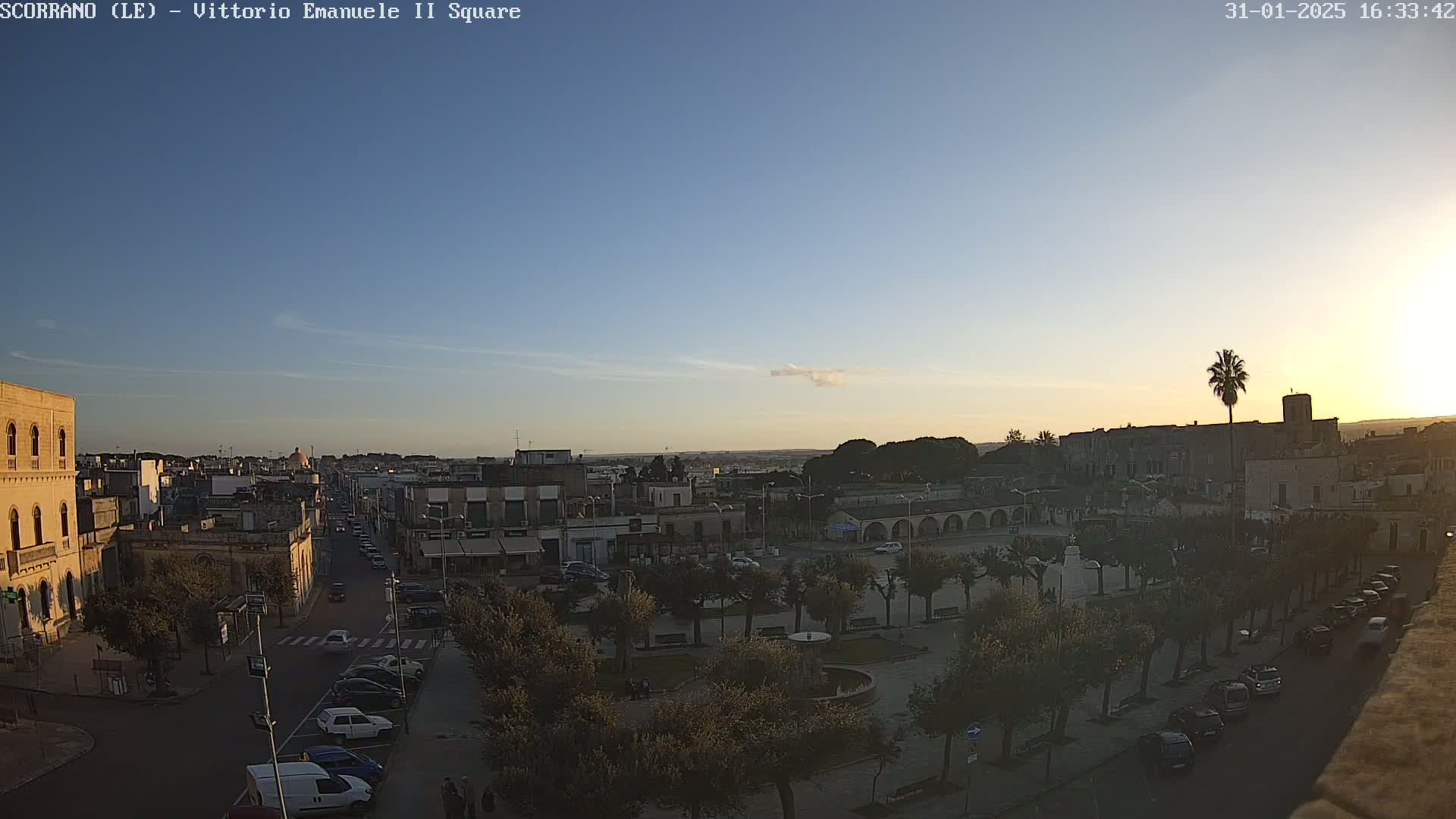 A high-angle view of a town square at sunset, under a clear sky with a few wispy clouds, shows cars parked along streets lined with trees, and buildings in the background.