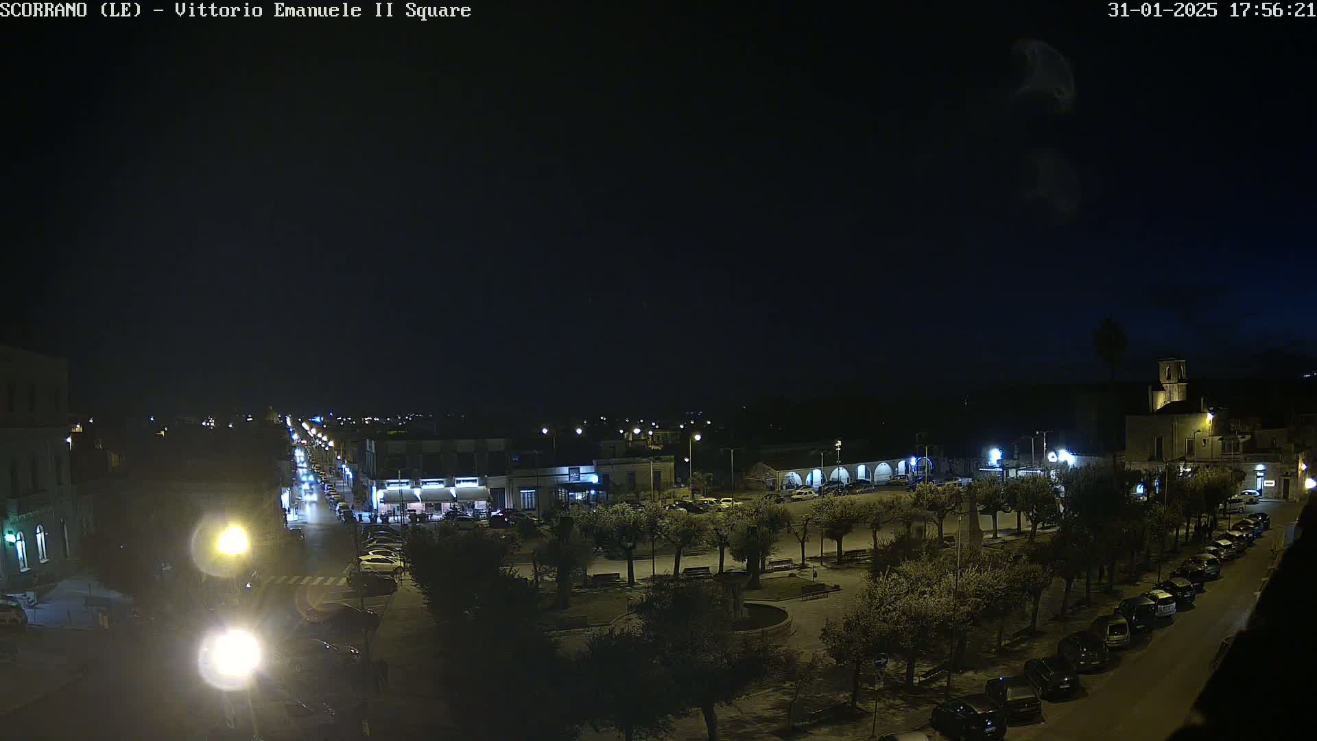 A nighttime, high-angle view of a town square with parked cars lining the streets and trees illuminated by streetlights.