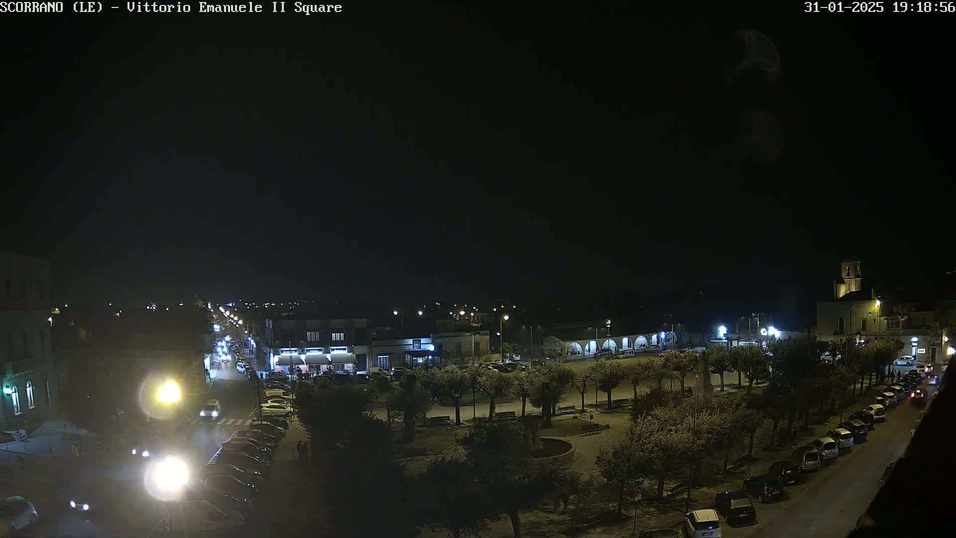 A nighttime, overhead view of a town square with parked cars lining the streets and trees in the center, under a clear, dark sky.