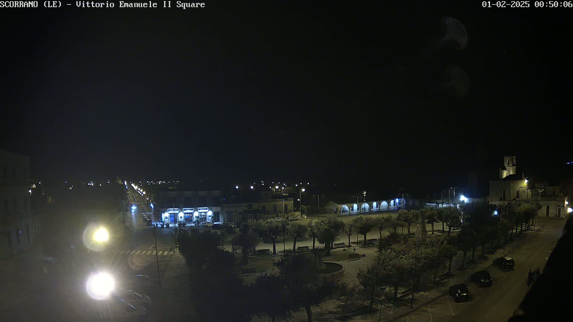 A nighttime view of a town square, illuminated by streetlights, with a few cars parked on the streets and trees lining the square.