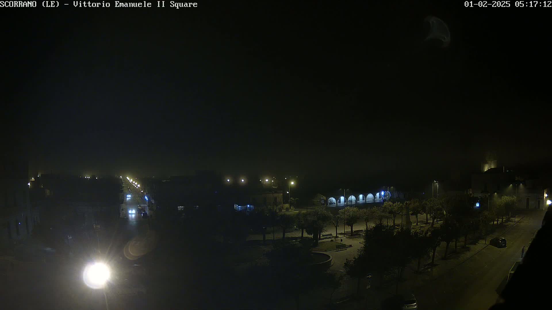 A nighttime, hazy view of a dimly lit town square with a street leading away from it, showing a few vehicles and trees.