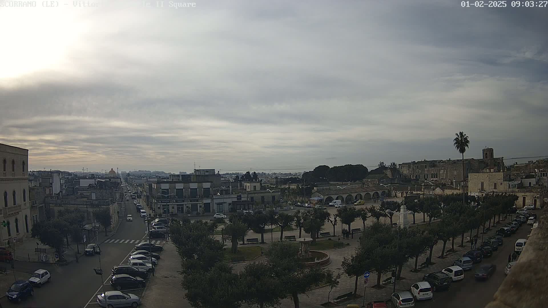 A mostly cloudy day reveals a town square with parked cars and trees, overlooked by buildings and a distant coastline.