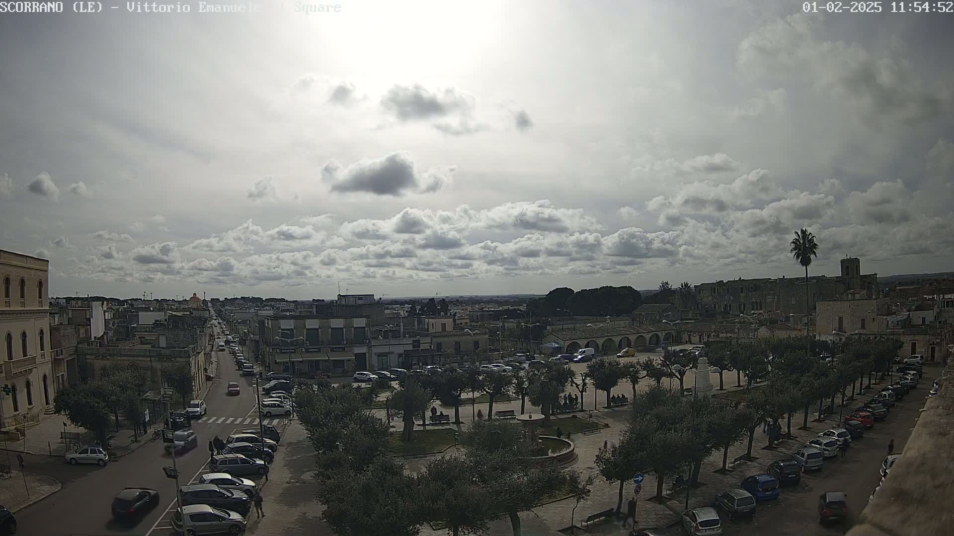 A partly cloudy day reveals a town square with parked cars, a fountain, and numerous trees lining the streets, extending to buildings and a distant horizon.