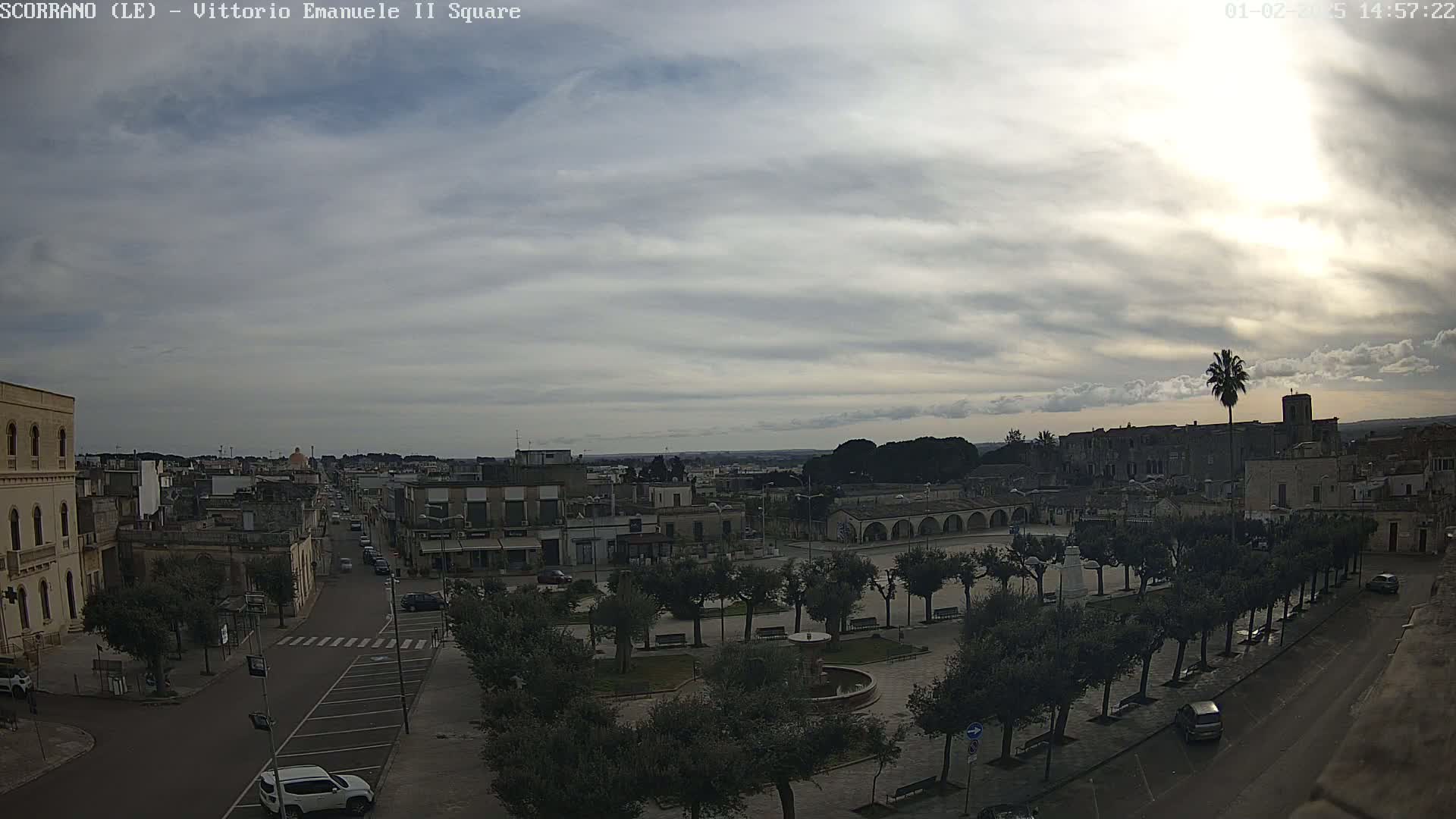 A mostly cloudy day reveals a town square with parked cars, trees lining a street, and buildings stretching into the distance.