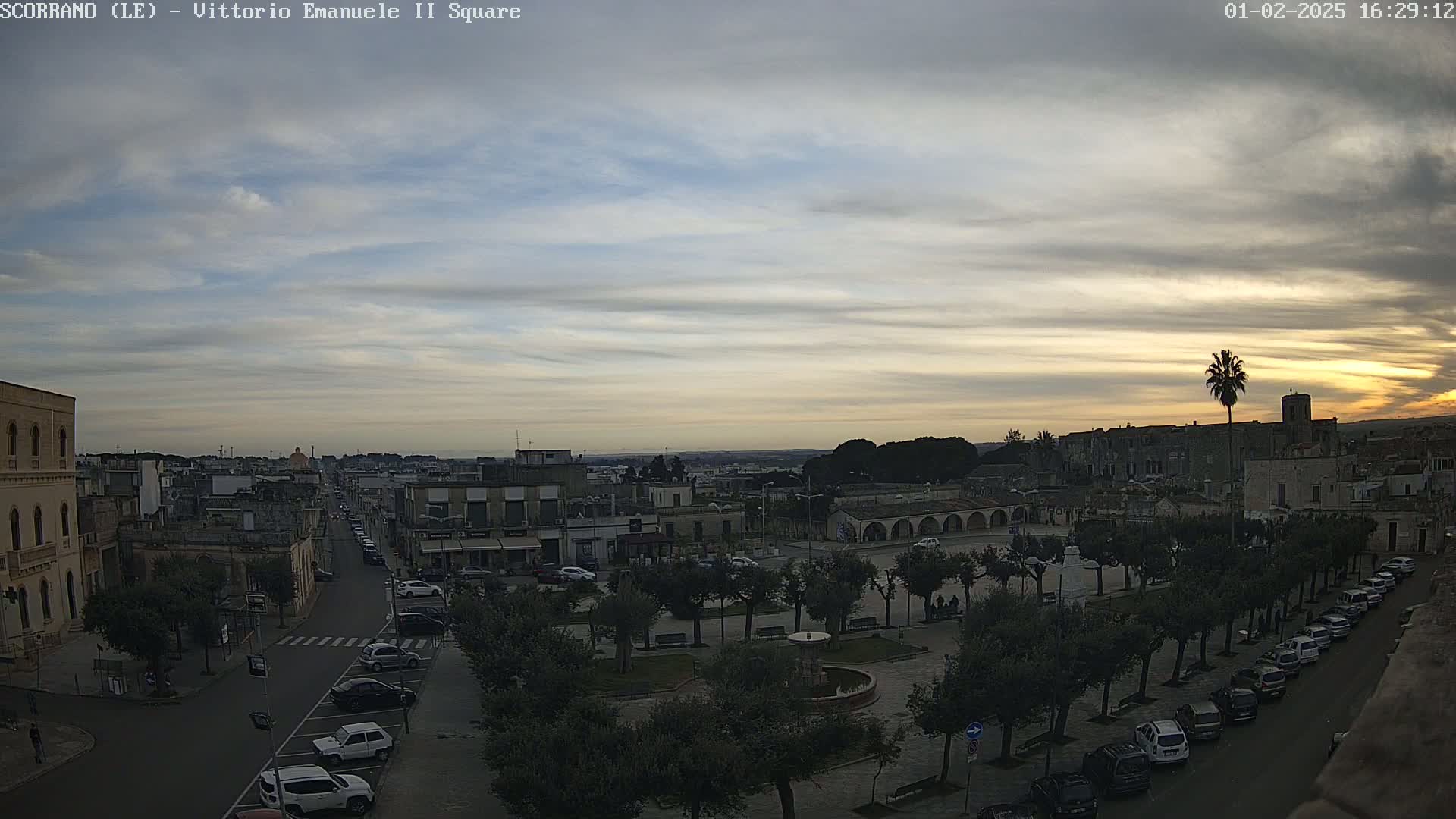 A high-angle view of a town square with parked cars lining the streets, under a mostly cloudy sunset sky.