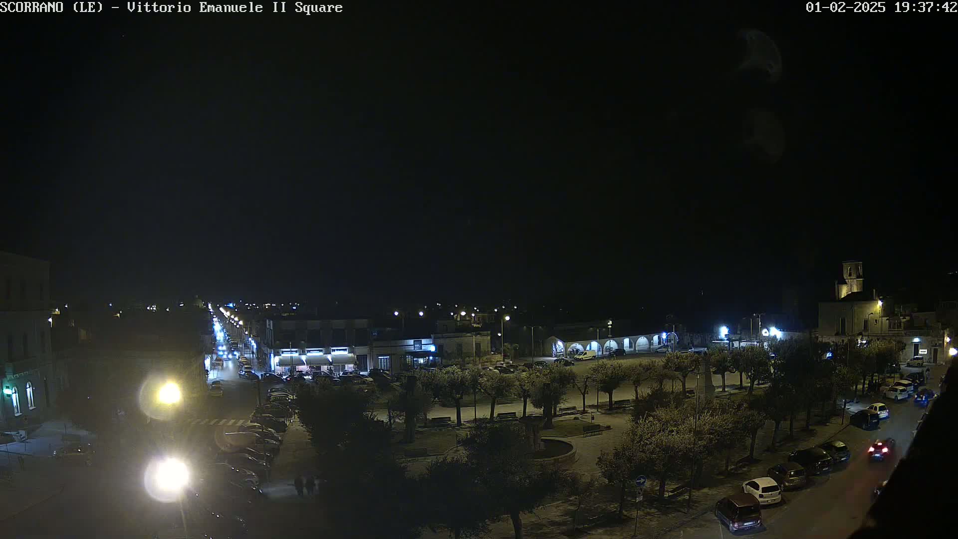 A nighttime view of a town square with cars parked along the sides, trees lining the perimeter, and streetlights illuminating the scene.