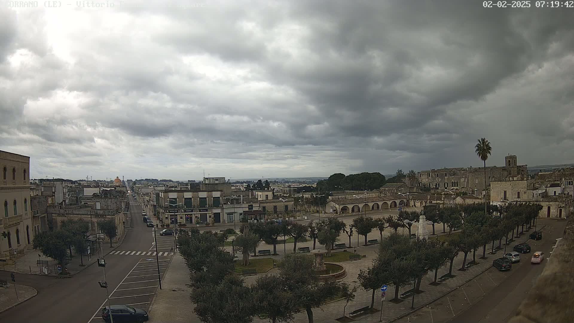 A town square with a fountain and several trees is visible under an overcast sky, with a street and buildings extending into the distance.