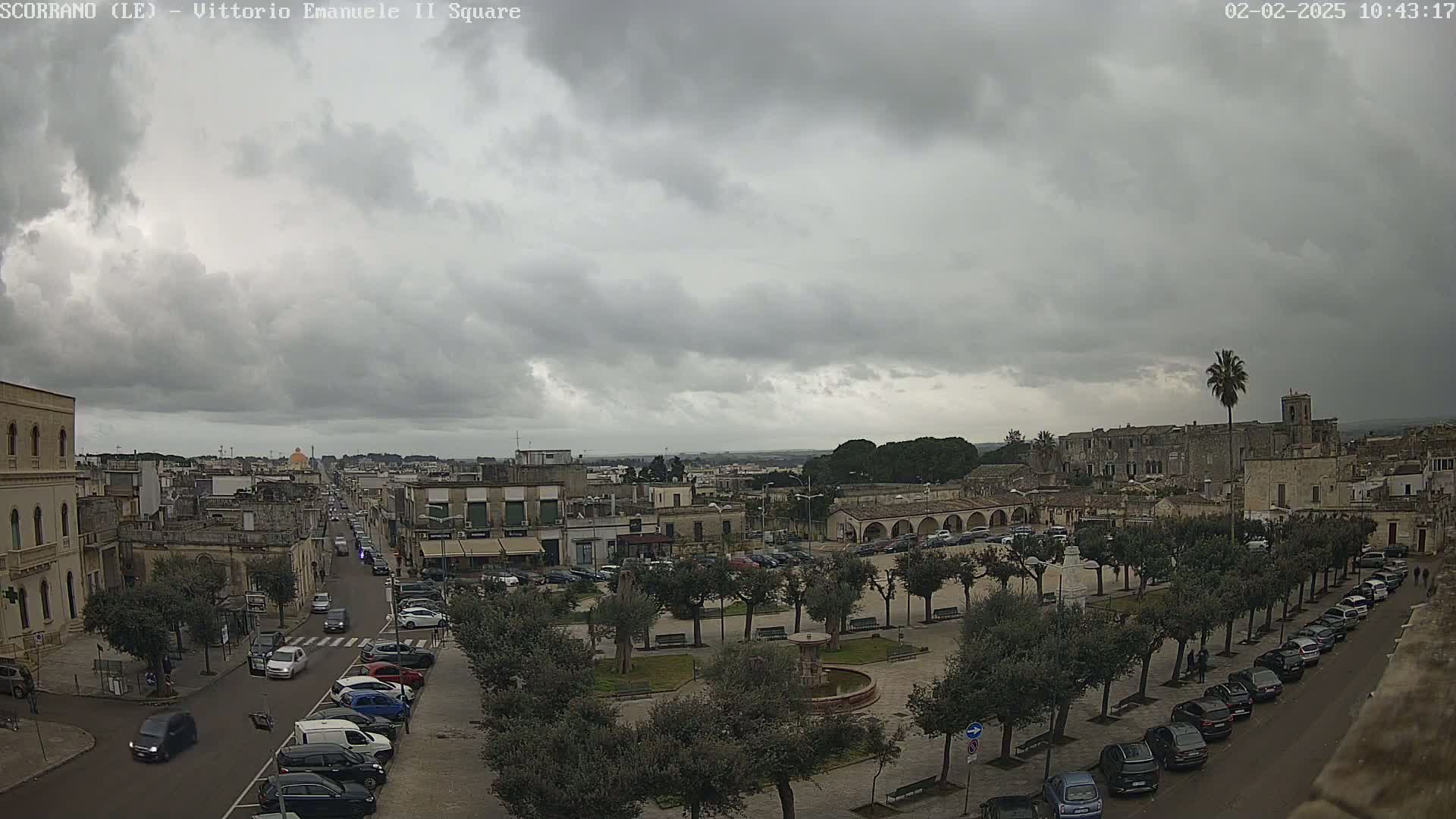 A town square with parked cars and a fountain is seen under a cloudy sky.