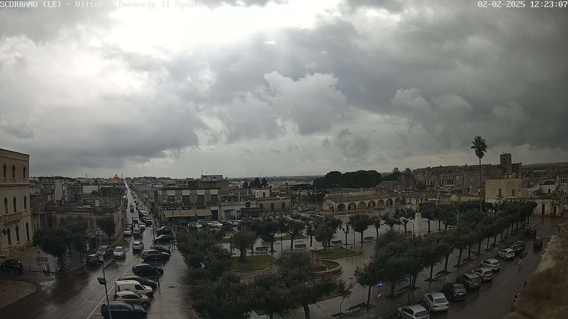 A wet town square with parked cars and a street with traffic under a cloudy, overcast sky.