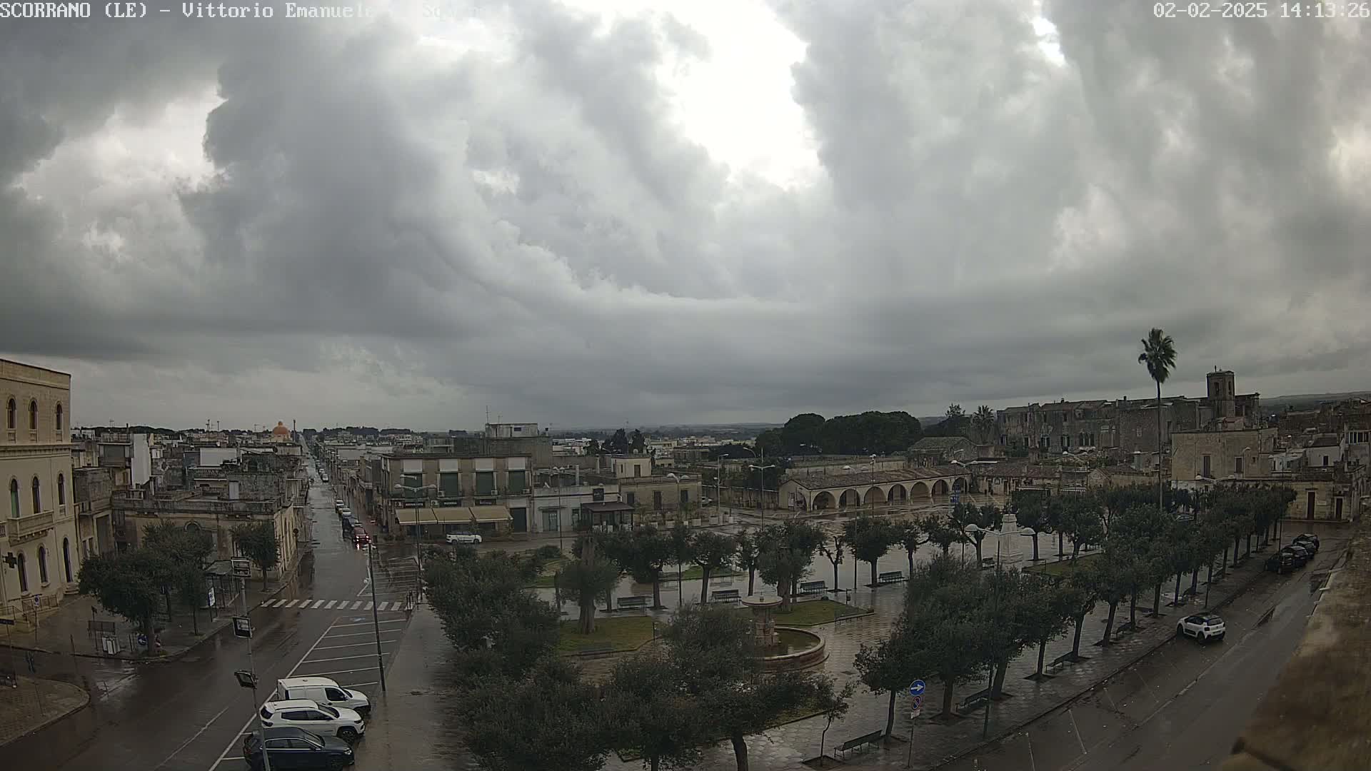 A wet town square with a fountain and rows of trees is seen under a cloudy sky, with buildings and streets extending into the distance.