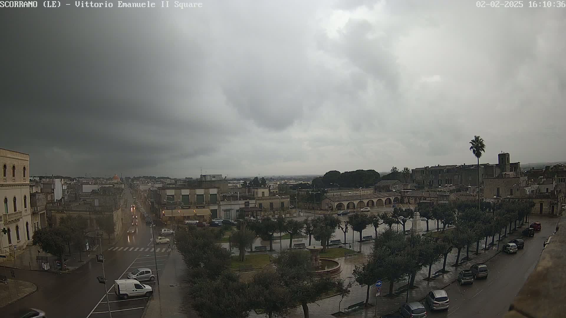 A wet town square is seen under a cloudy sky, with cars parked along the sides of a street and buildings surrounding the area.