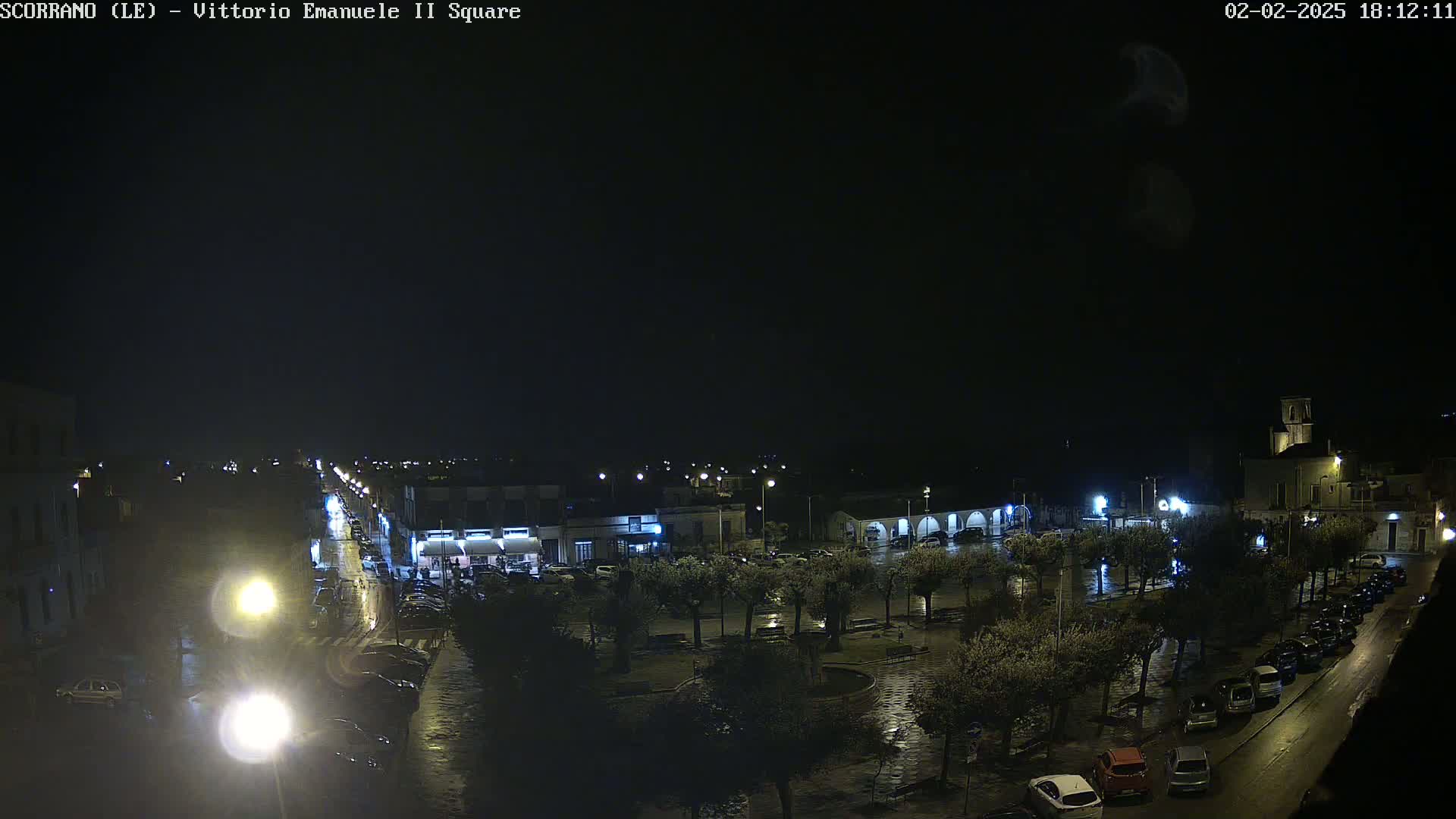 A wet, nighttime view of a town square with parked cars and streetlights illuminating the area.