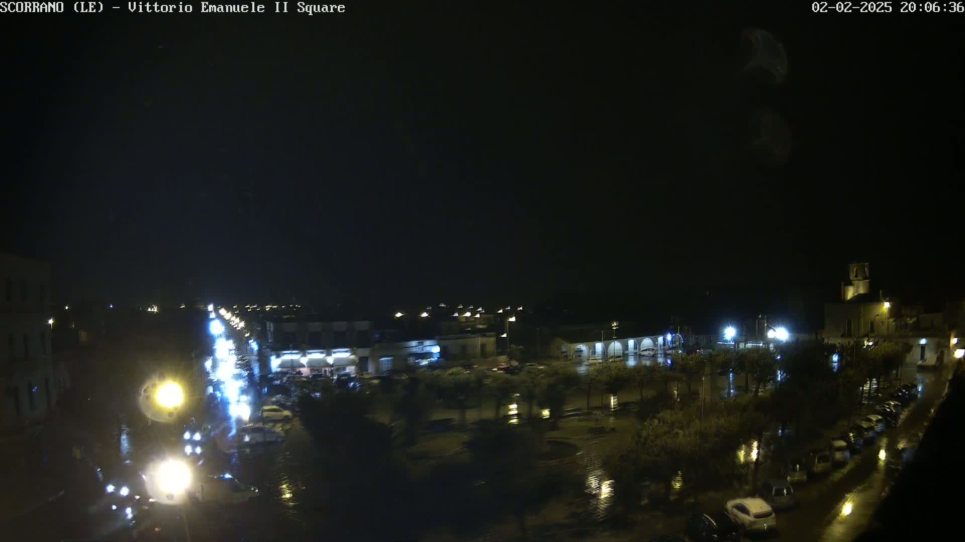 A nighttime view of a rain-soaked town square and street, with cars parked and driving under dim streetlights.