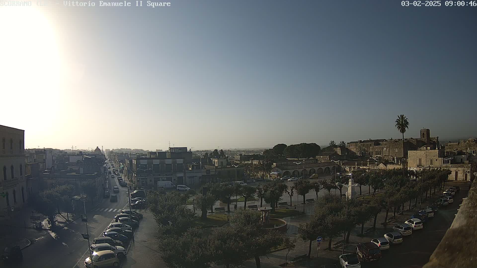 A high-angle, long shot of a town square with parked cars, trees, and buildings under a clear, bright sky.