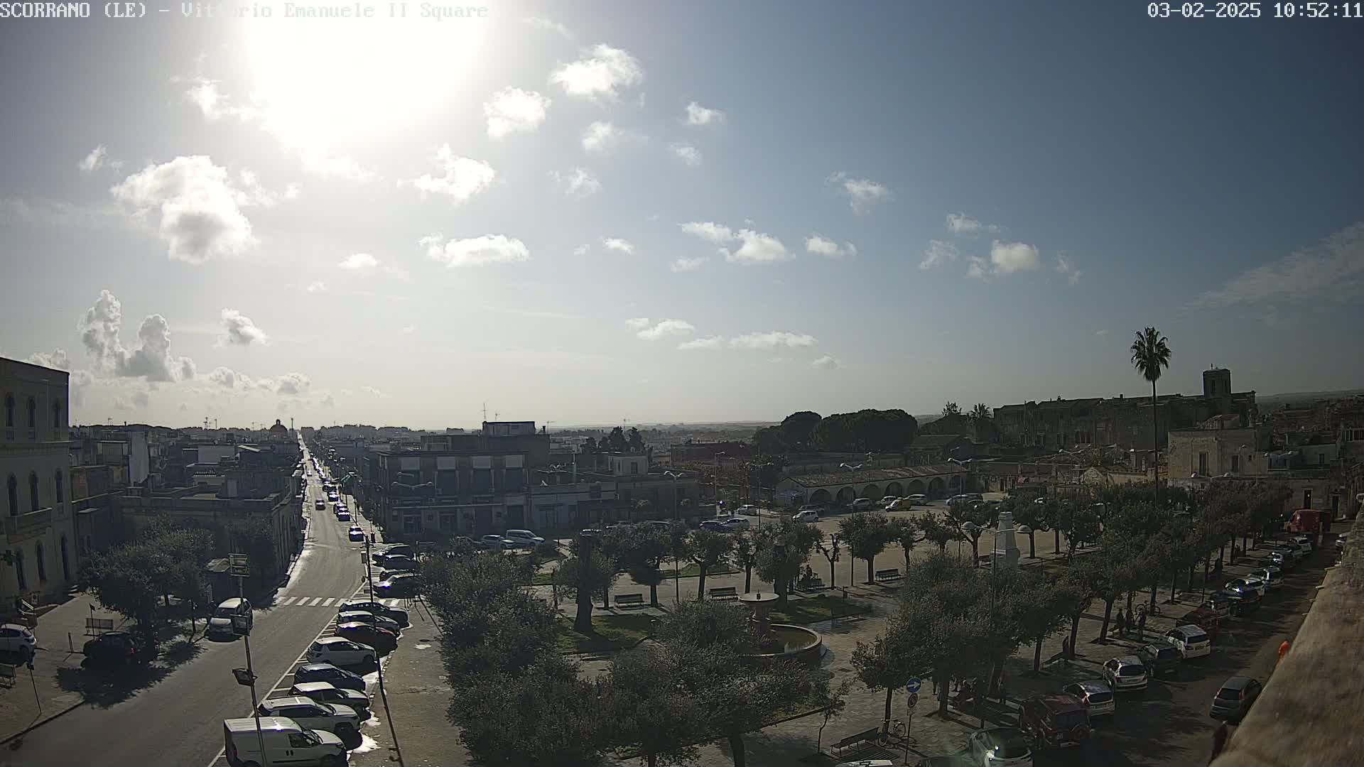 A partly sunny day reveals a town square with parked cars lining its edges, a fountain in the center, and a street leading to more buildings in the distance.