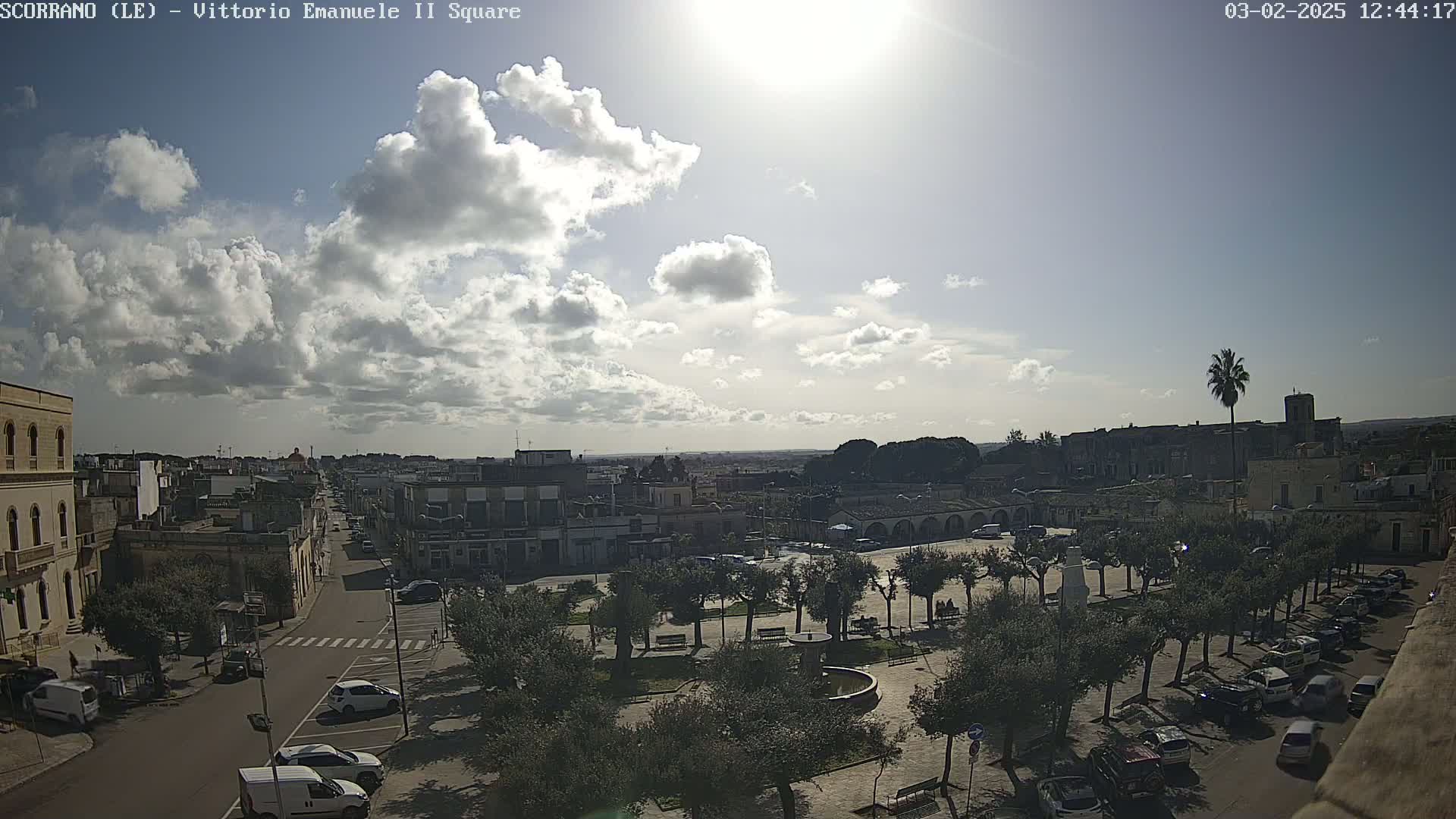 A partly sunny day reveals a town square with a fountain, surrounded by trees and parked cars, extending to buildings and streets beyond.