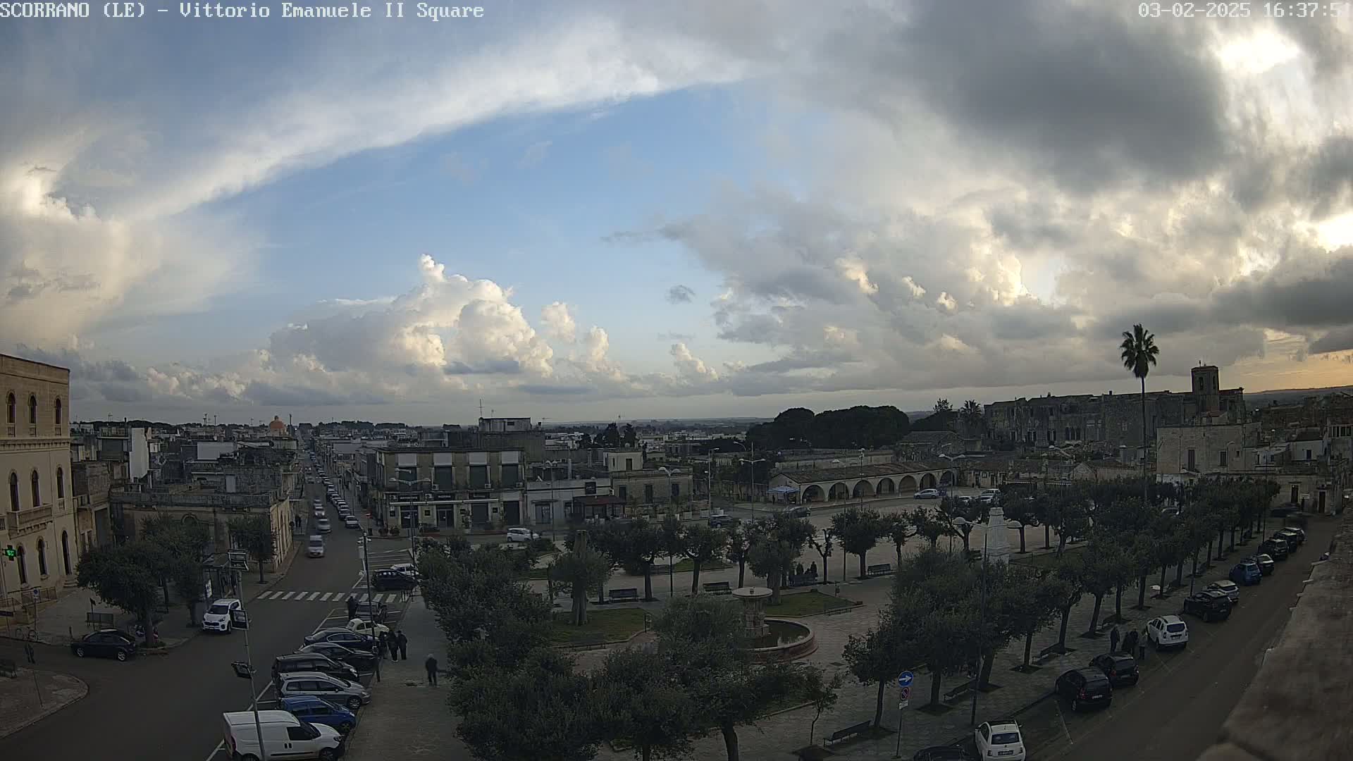 A partly cloudy sky overlooks a town square with parked cars and a street lined with vehicles, under a mostly cloudy sky.