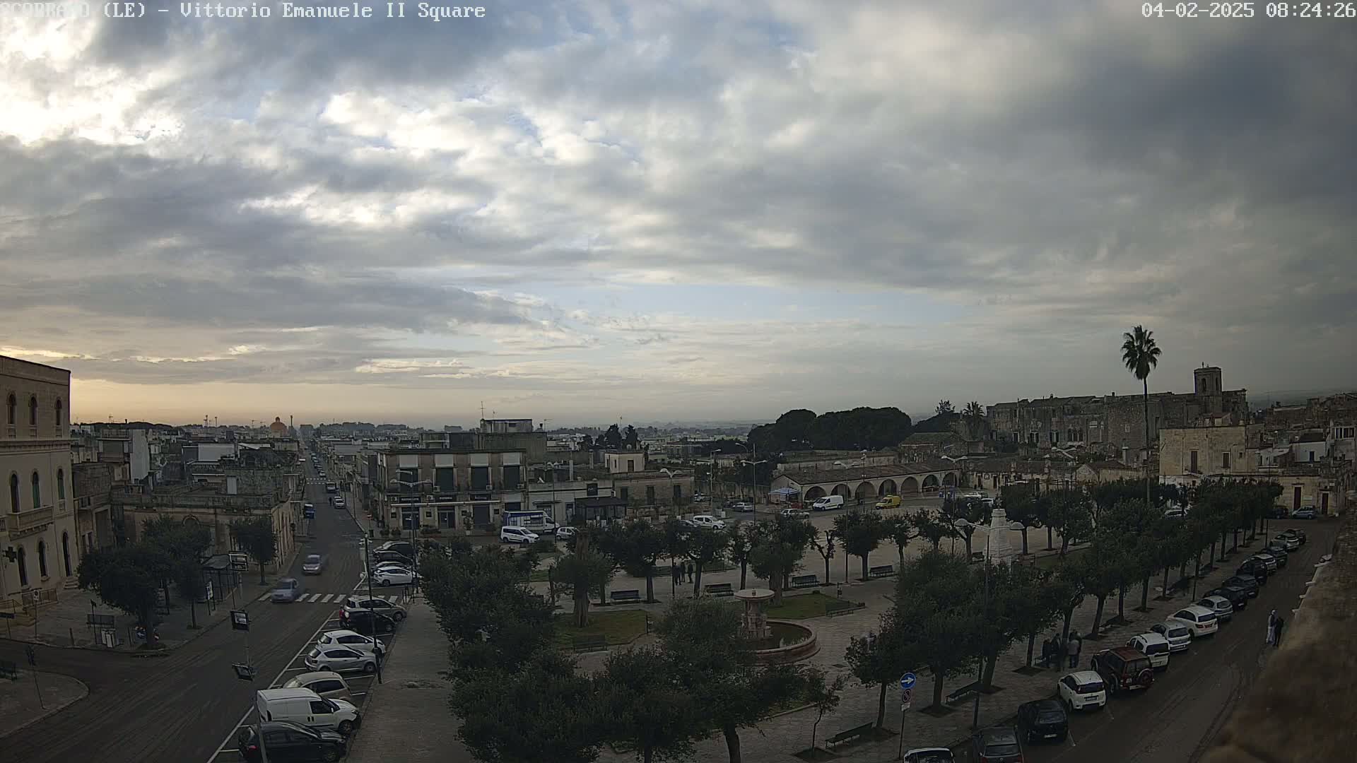 An overcast sky hangs over a town square with parked cars lining the streets, and a fountain in the center of the square, under a cloudy sky.