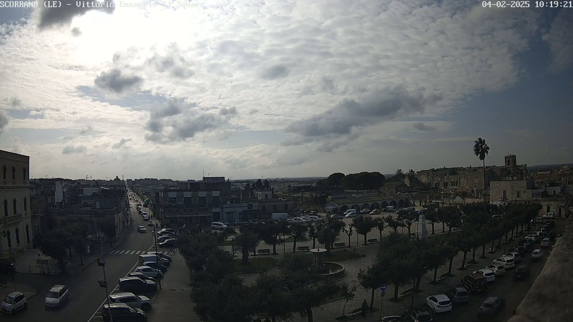 A partly cloudy day reveals a town square with parked cars and lined trees, a street with cars driving, and buildings extending into the distance.