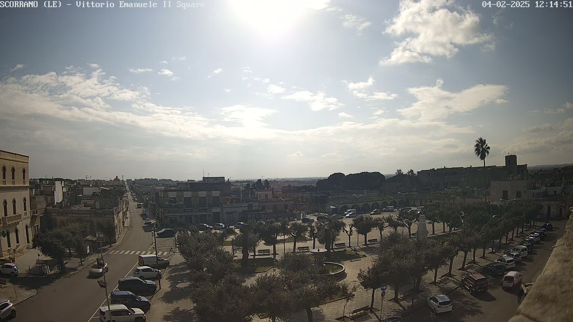 A sunny day reveals a town square with parked cars and olive trees, overlooked by buildings and a street leading to a wider cityscape under a partly cloudy sky.