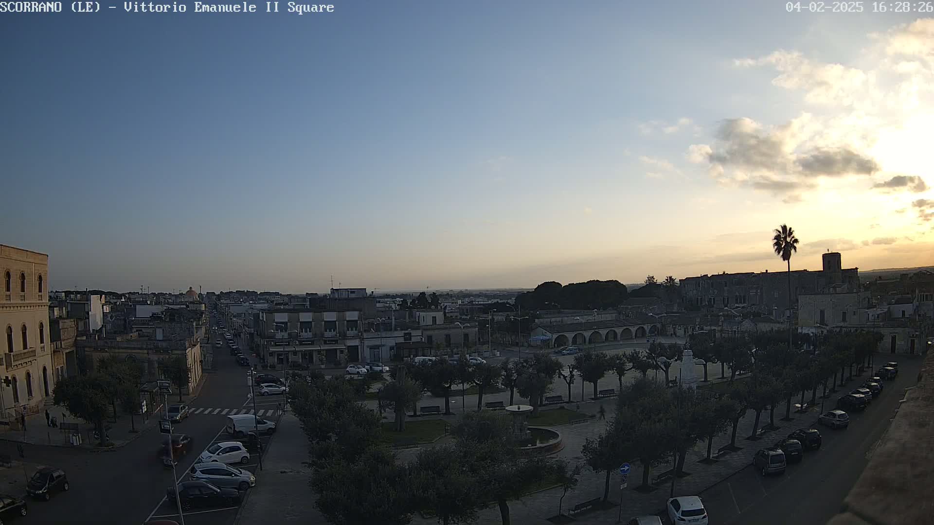 An aerial view of a town square, under a partly cloudy sunset sky, shows parked cars lining streets leading to the square, which features trees, a fountain, and buildings surrounding it.