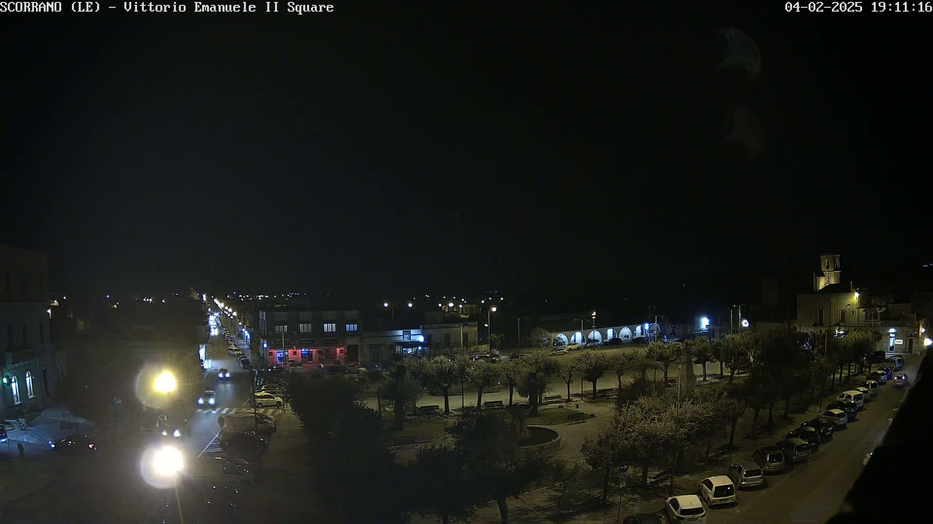 A nighttime view of a town square with parked cars lining the streets and a few vehicles on the road, under a dark, clear sky.