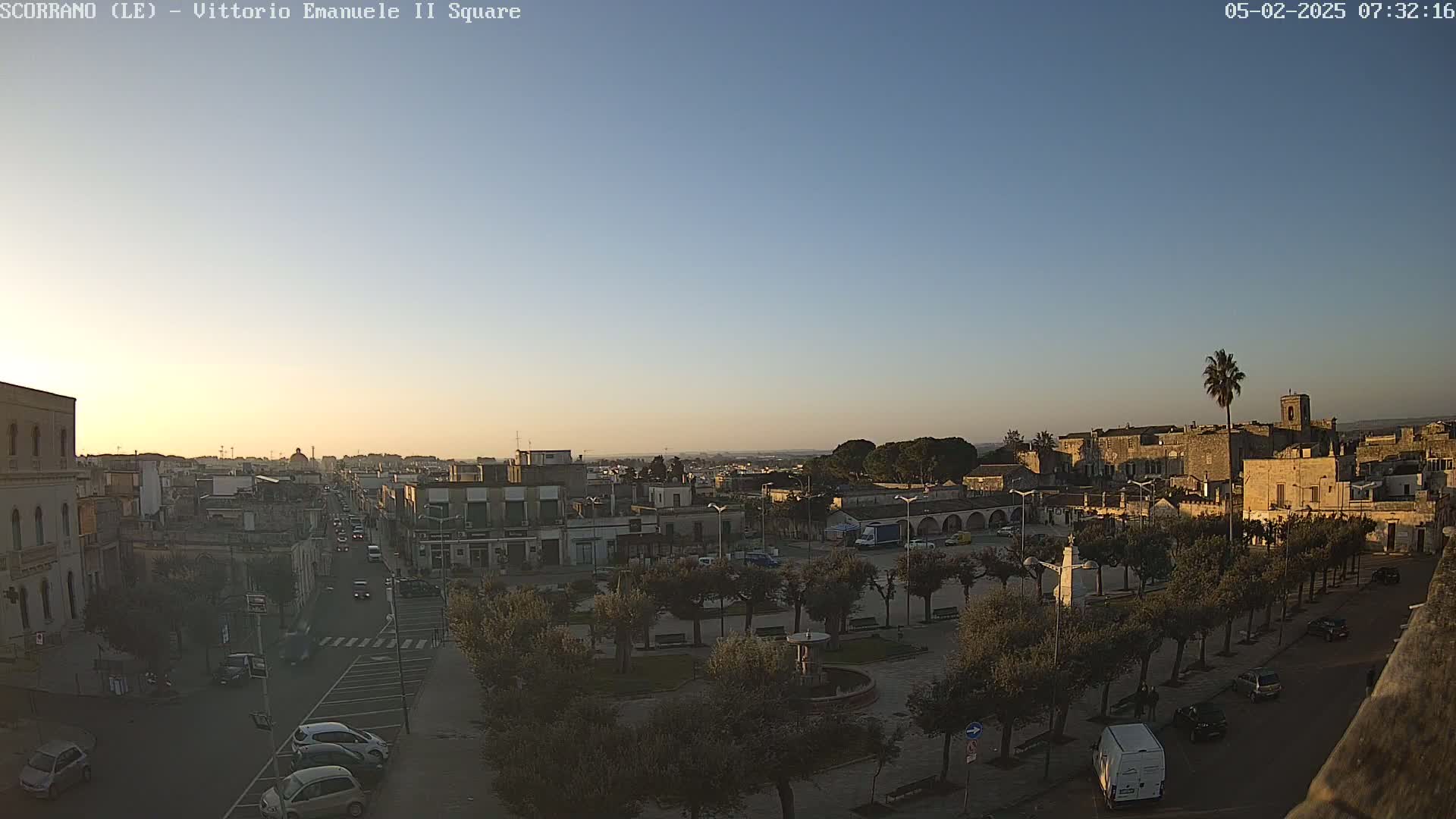 A town square with a fountain and rows of trees is visible at sunrise under a clear sky, with streets and buildings surrounding the square and cars parked along the sides of the road.