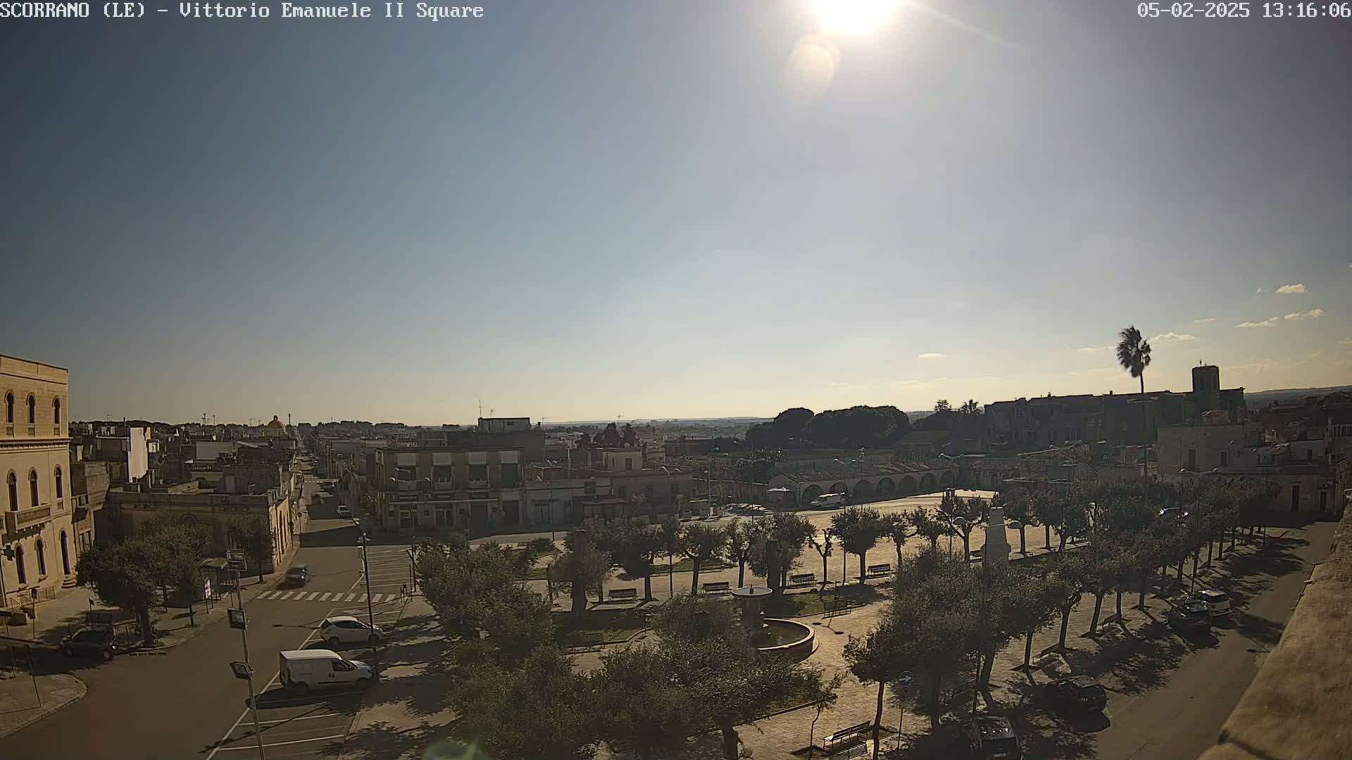A sunny day reveals a town square with a central fountain, surrounded by trees and buildings, and a few cars parked nearby.
