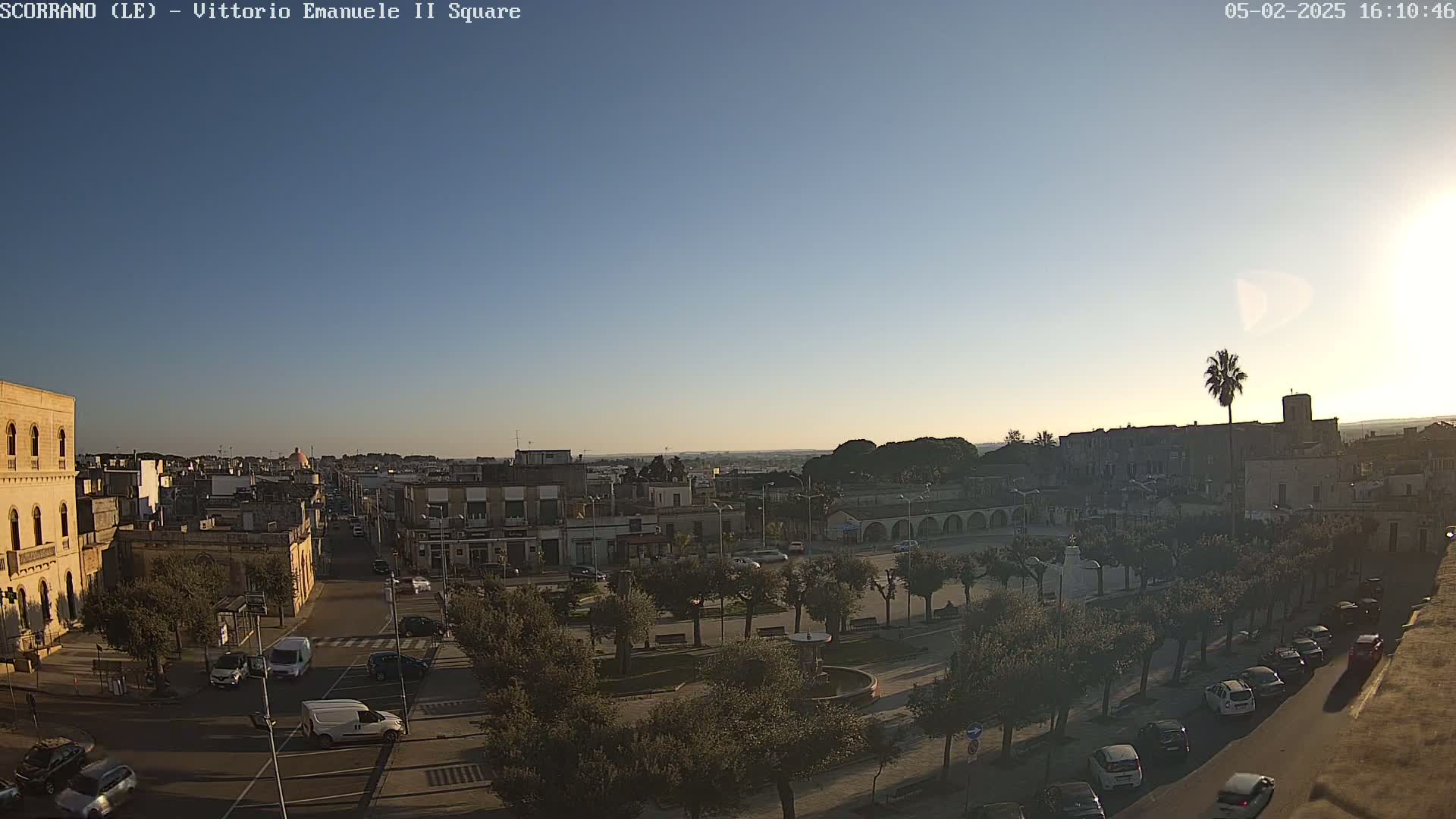 A clear, sunny day reveals a town square with parked cars lining the streets, trees, and buildings in the background.