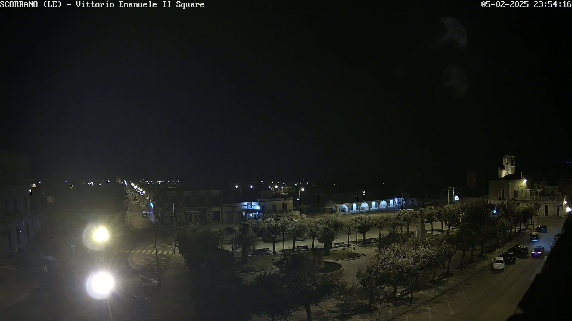 A nighttime view of a town square with trees, buildings, and a few cars, under a dark, clear sky.