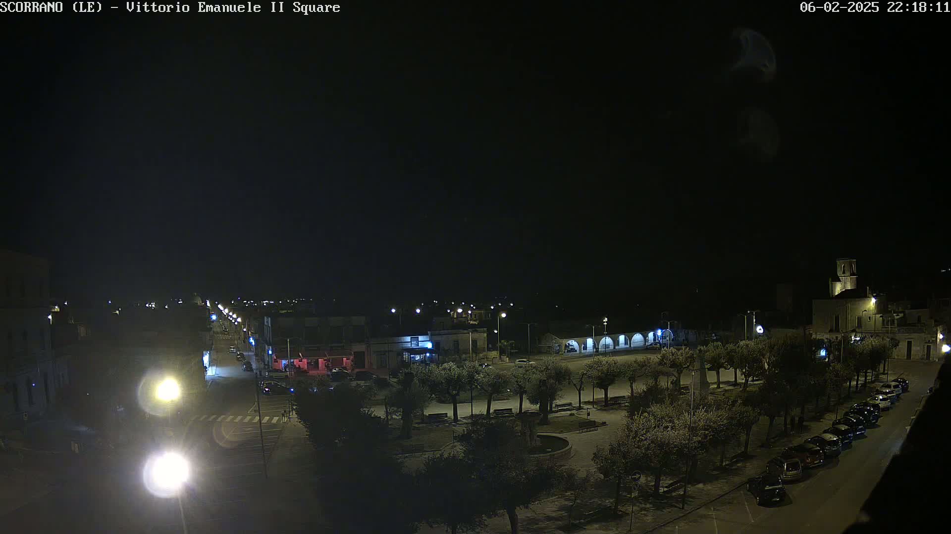 A nighttime, high-angle view of a town square with cars parked along the sides, illuminated by streetlights and surrounded by trees.