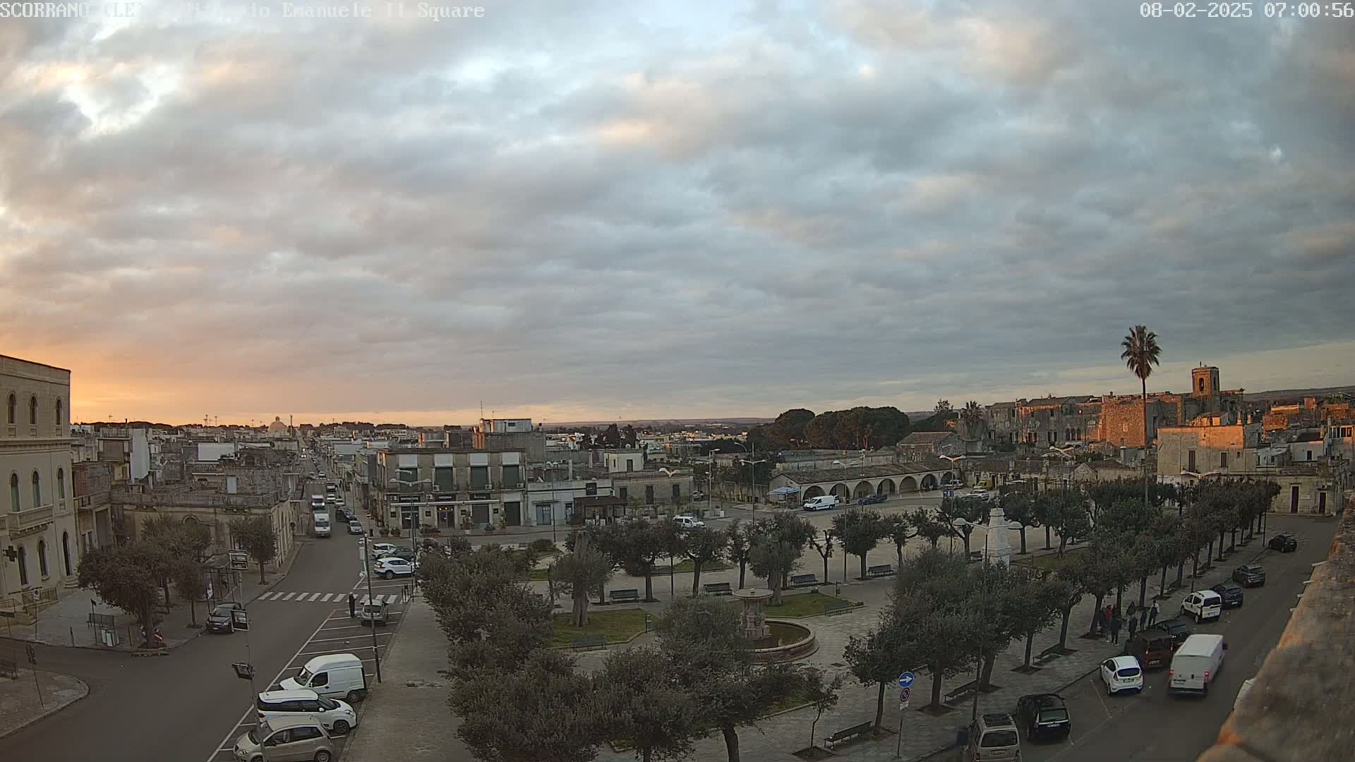 An overcast sunrise illuminates a town square with cars parked along its edges and streets, surrounded by low buildings and trees.