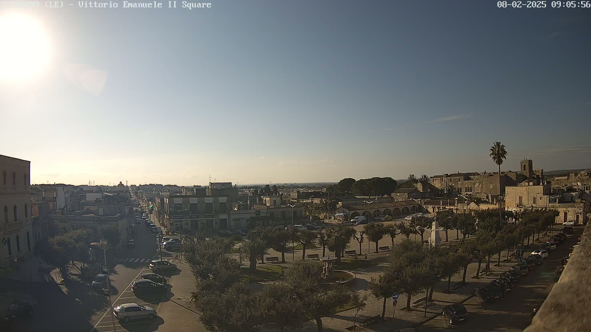 A sunny, clear day reveals a town square with a central fountain, surrounded by parked cars and olive trees, overlooking a cityscape extending to a distant horizon.