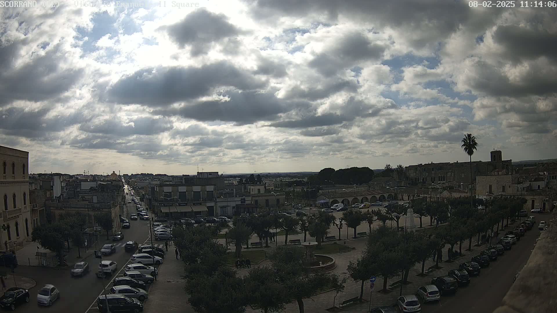 A partly cloudy day overlooks a town square with parked cars lining the streets and a fountain in the center, surrounded by trees.