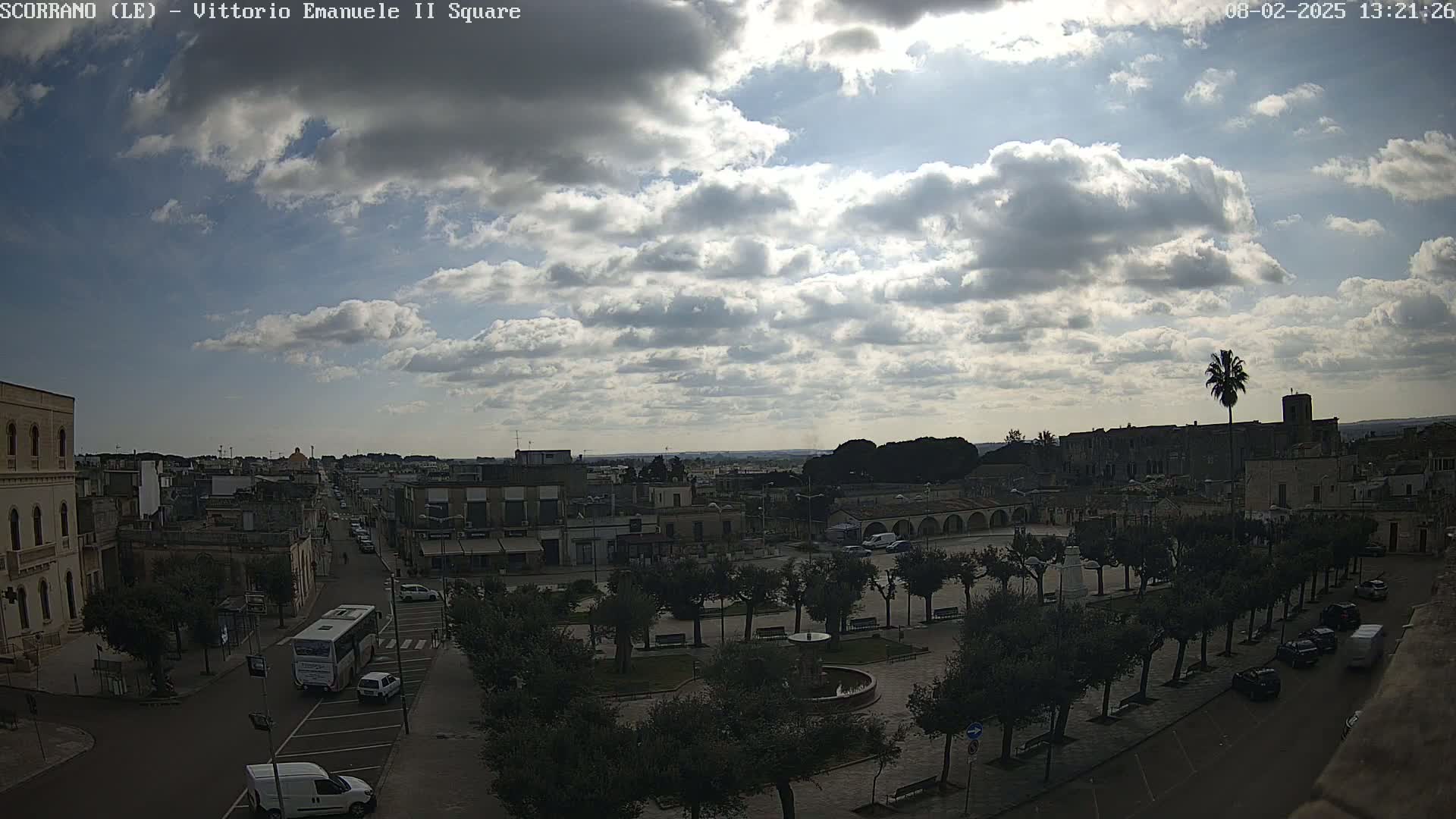 A partly cloudy sky overlooks a town square with trees, buildings, vehicles, and a fountain, under what appears to be bright, but diffused sunlight.