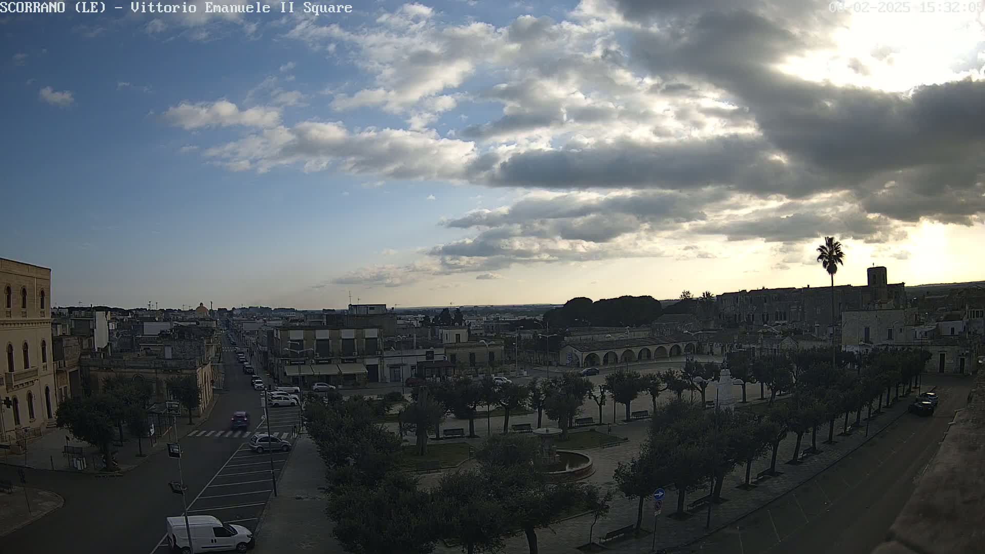 A partly cloudy sunset over a town square with a fountain, lined with trees, and a street with cars.