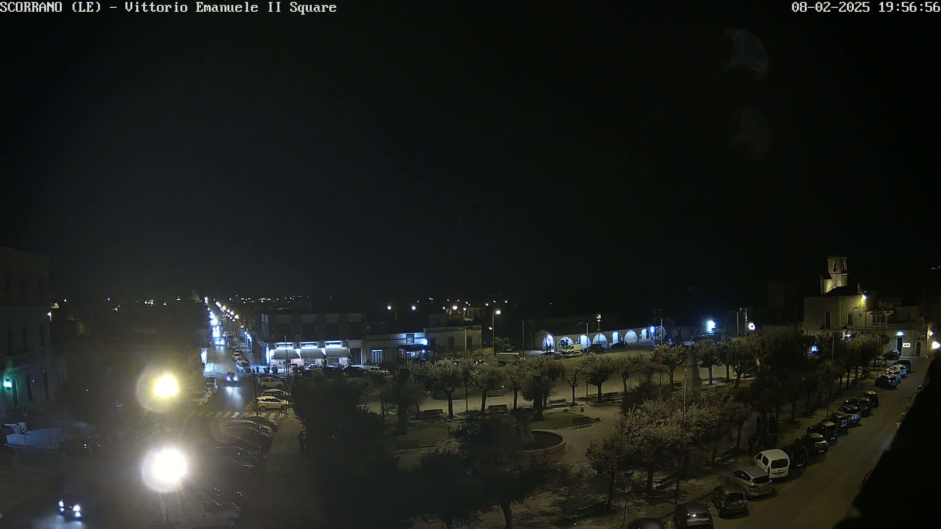 A nighttime, high-angle view of a town square with parked cars lining the streets, illuminated by streetlights, and trees sparsely scattered within the square.