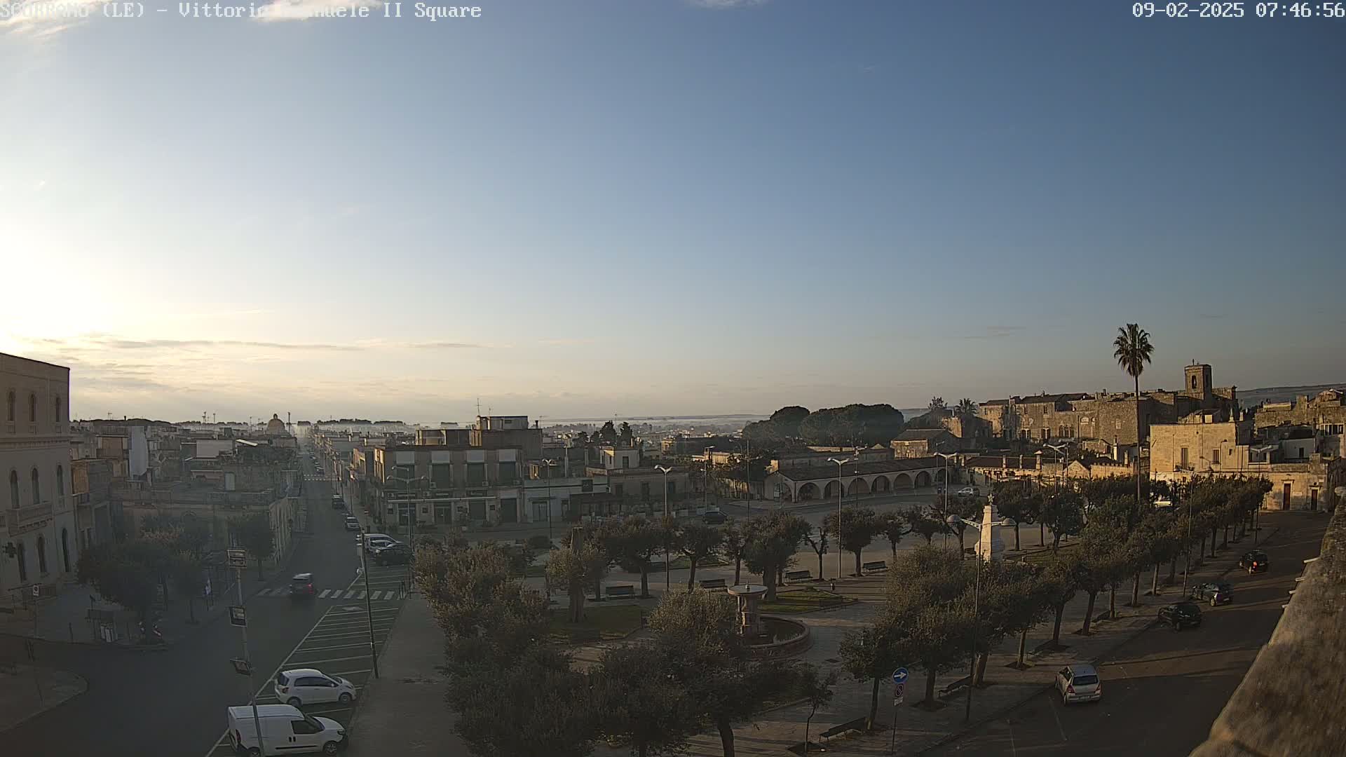 A town square with a fountain and trees is visible, with a clear sky and distant buildings under bright, early morning sunlight.