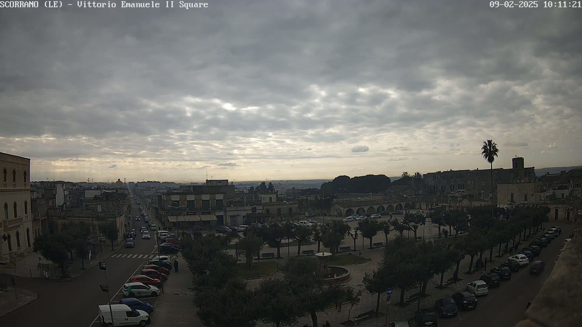 A city square with parked cars and a fountain is viewed from above under a cloudy sky.
