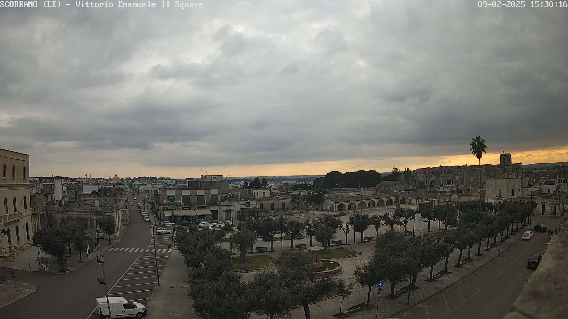 A mostly cloudy sunset over a town square with a fountain and rows of trees, viewed from an elevated position.