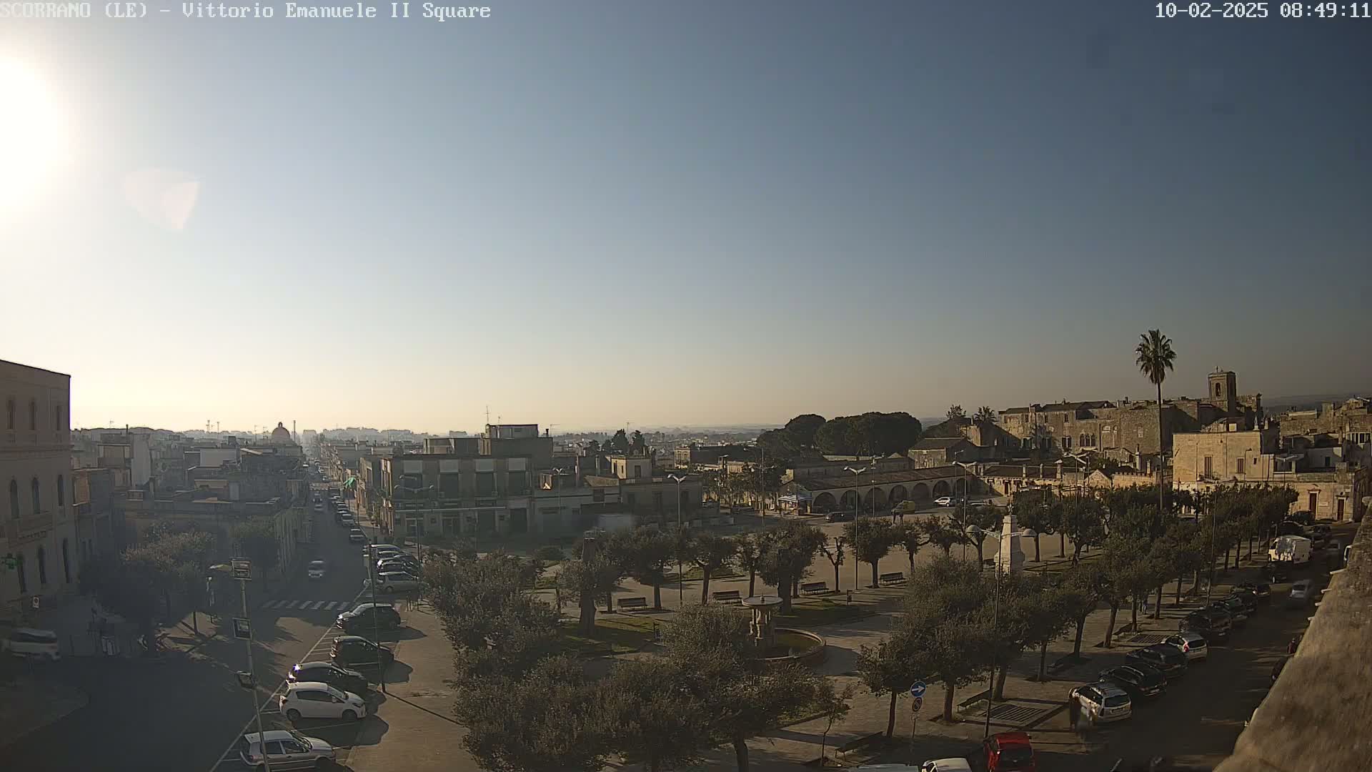 A high-angle, long shot shows a town square with parked cars and a few pedestrians under a clear, bright sky.