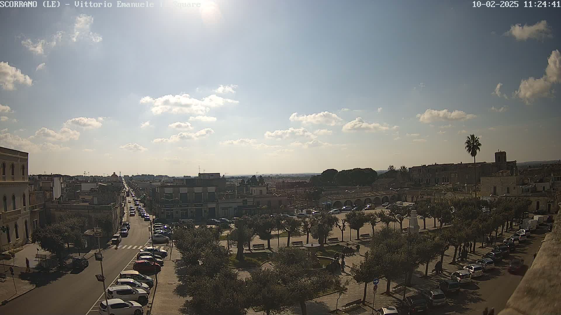 A partly sunny day reveals a town square with parked cars lining the streets, surrounded by buildings and olive trees under a mostly clear sky.
