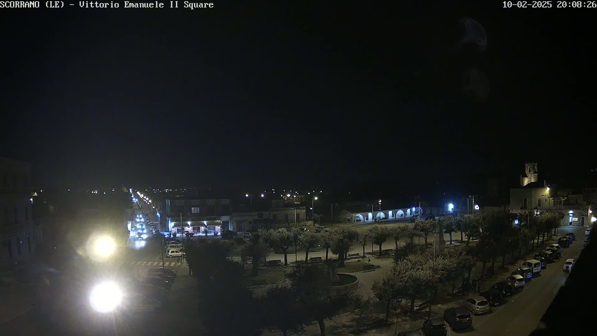 A nighttime, overhead view of a town square with cars parked along the street and trees lining the perimeter of the square, under a clear, dark sky.