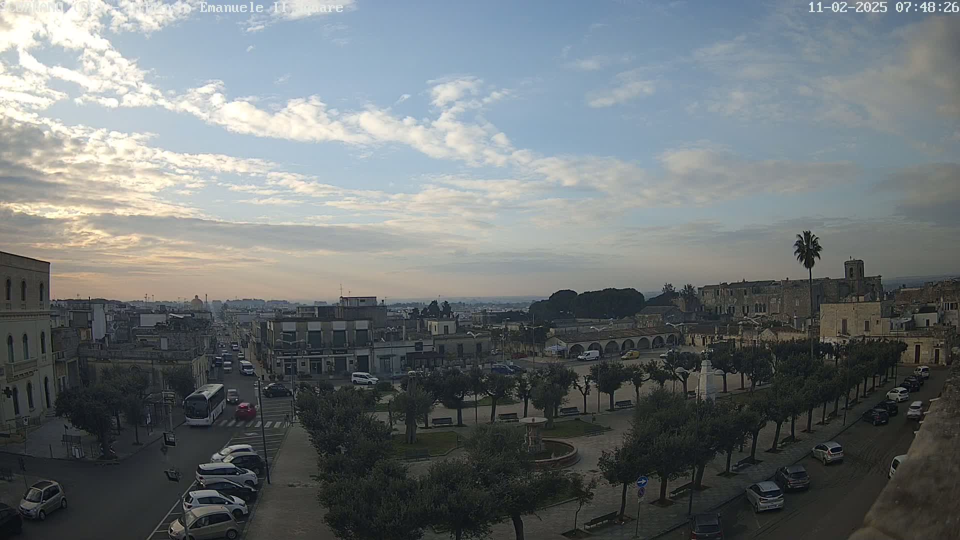 A partly cloudy sky overlooks a town square with parked cars and trees, and a city extending into the distance.