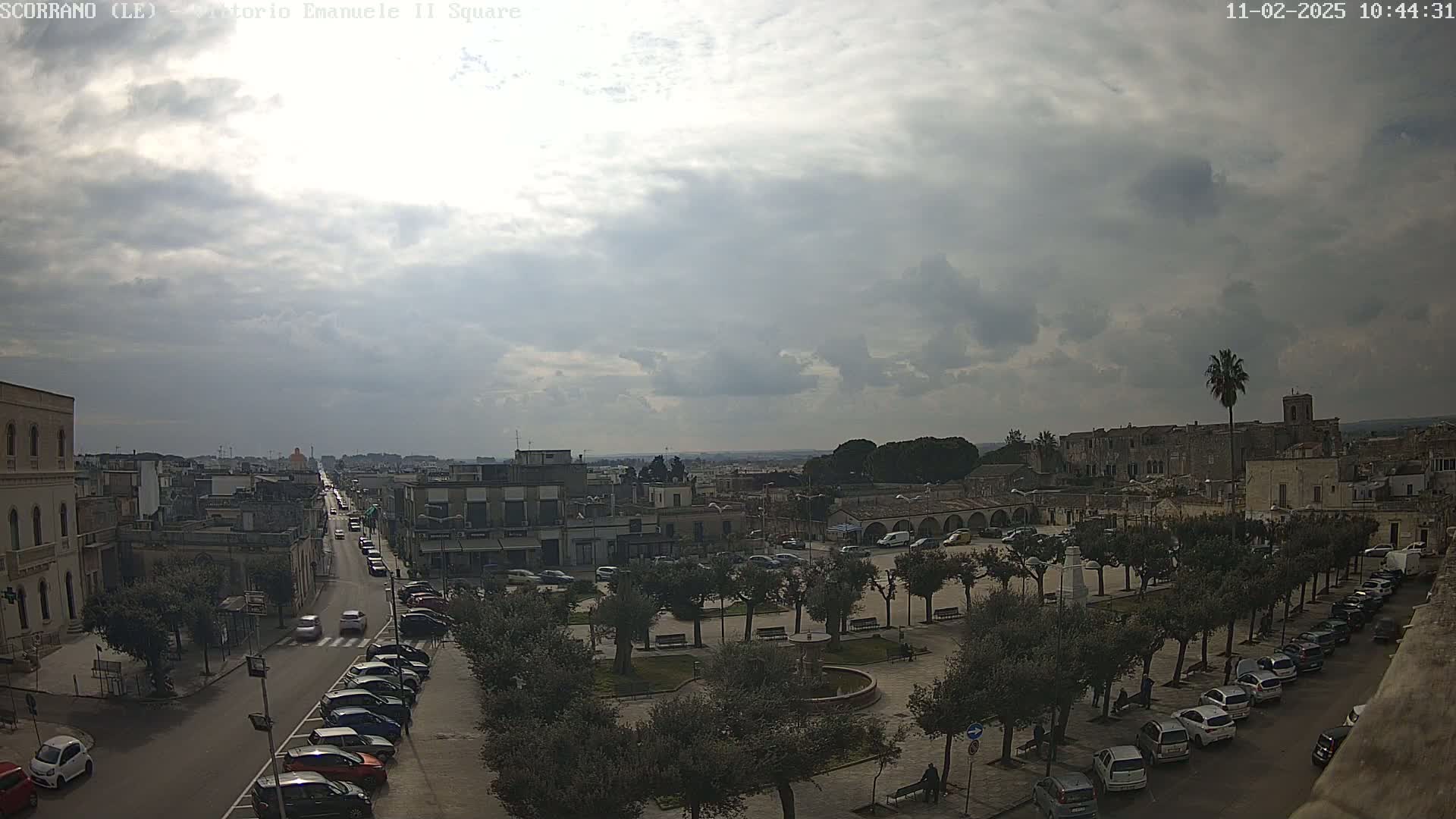 A mostly cloudy day overlooks a town square with parked cars lining the streets and a fountain in the center, surrounded by trees.