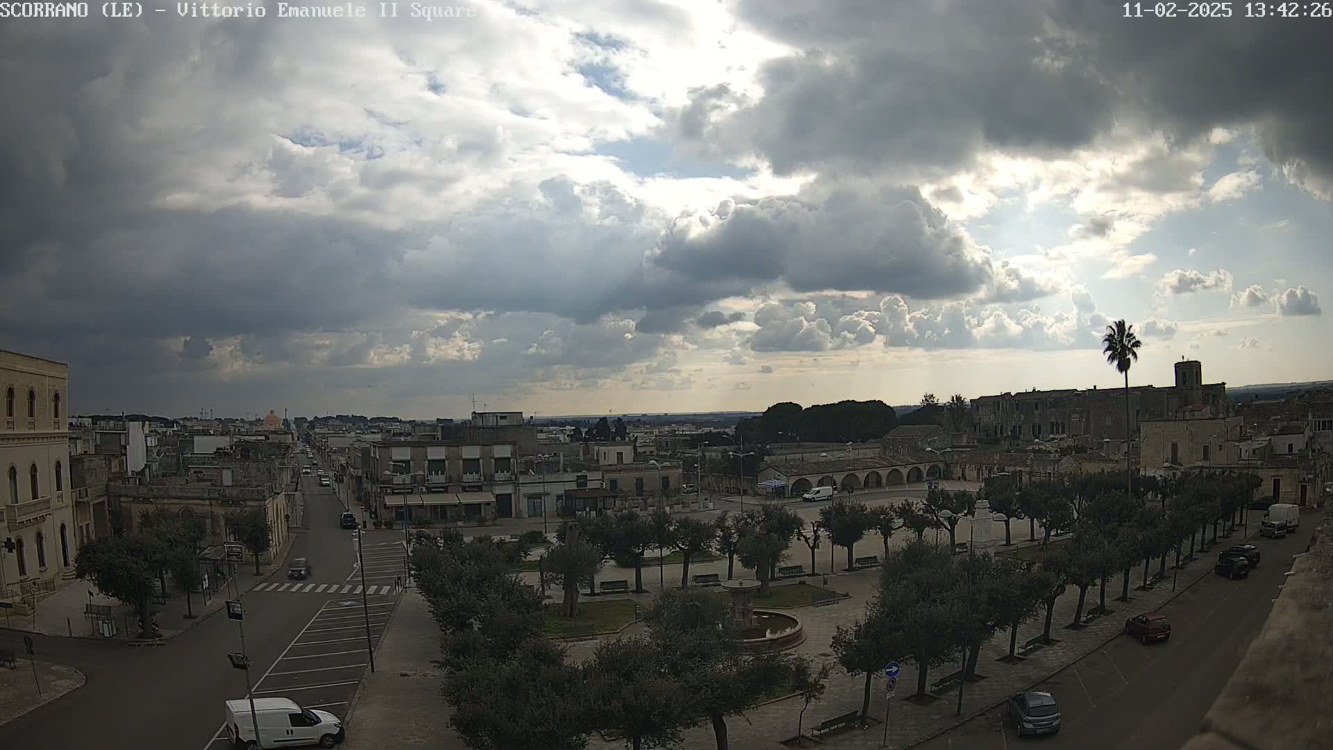 A partly cloudy sky overlooks a town square with a fountain and trees, and a street lined with cars leading to other buildings.