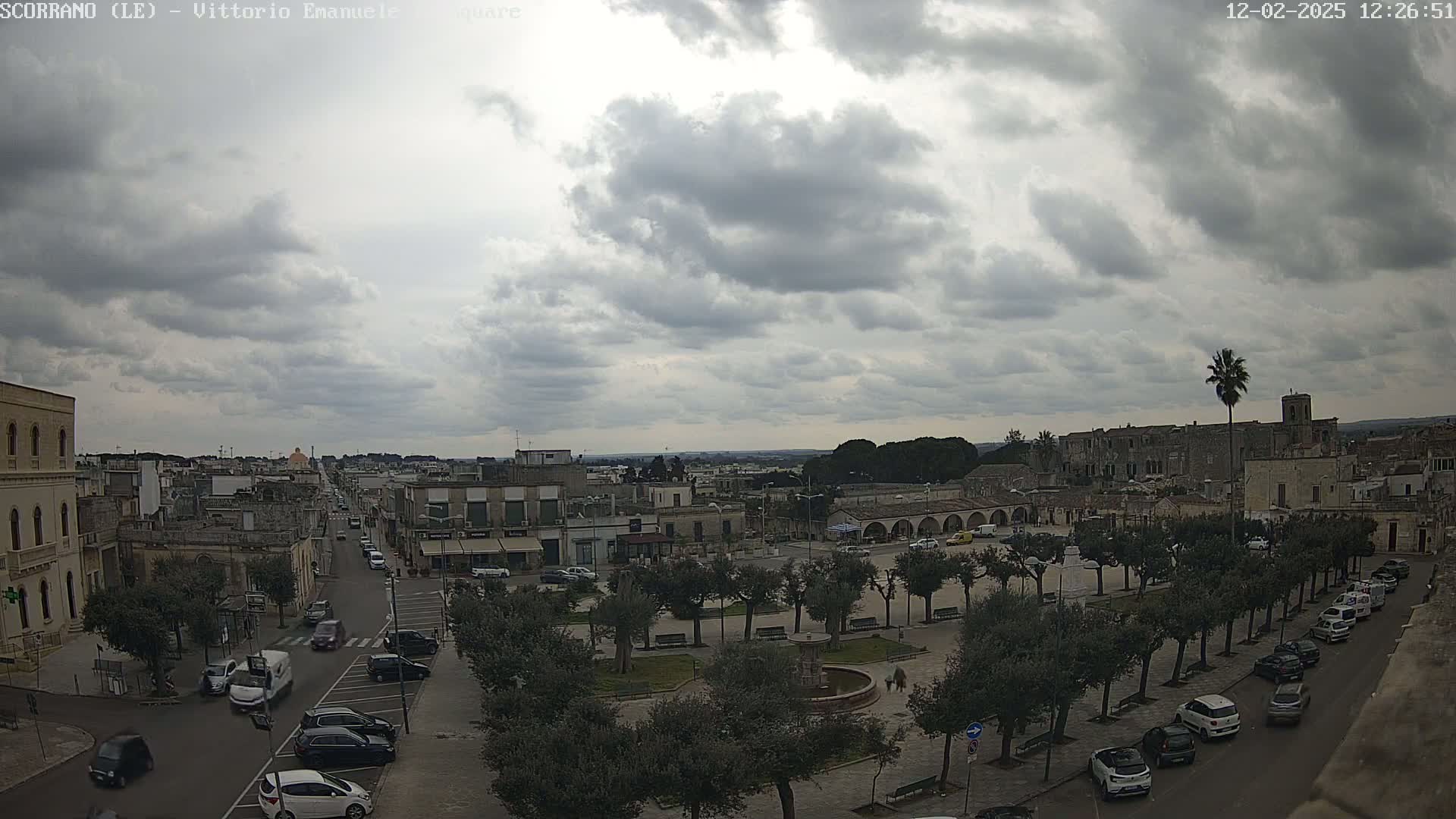 An overcast sky hangs over a town square, where cars are parked along tree-lined streets and buildings surround a central fountain.