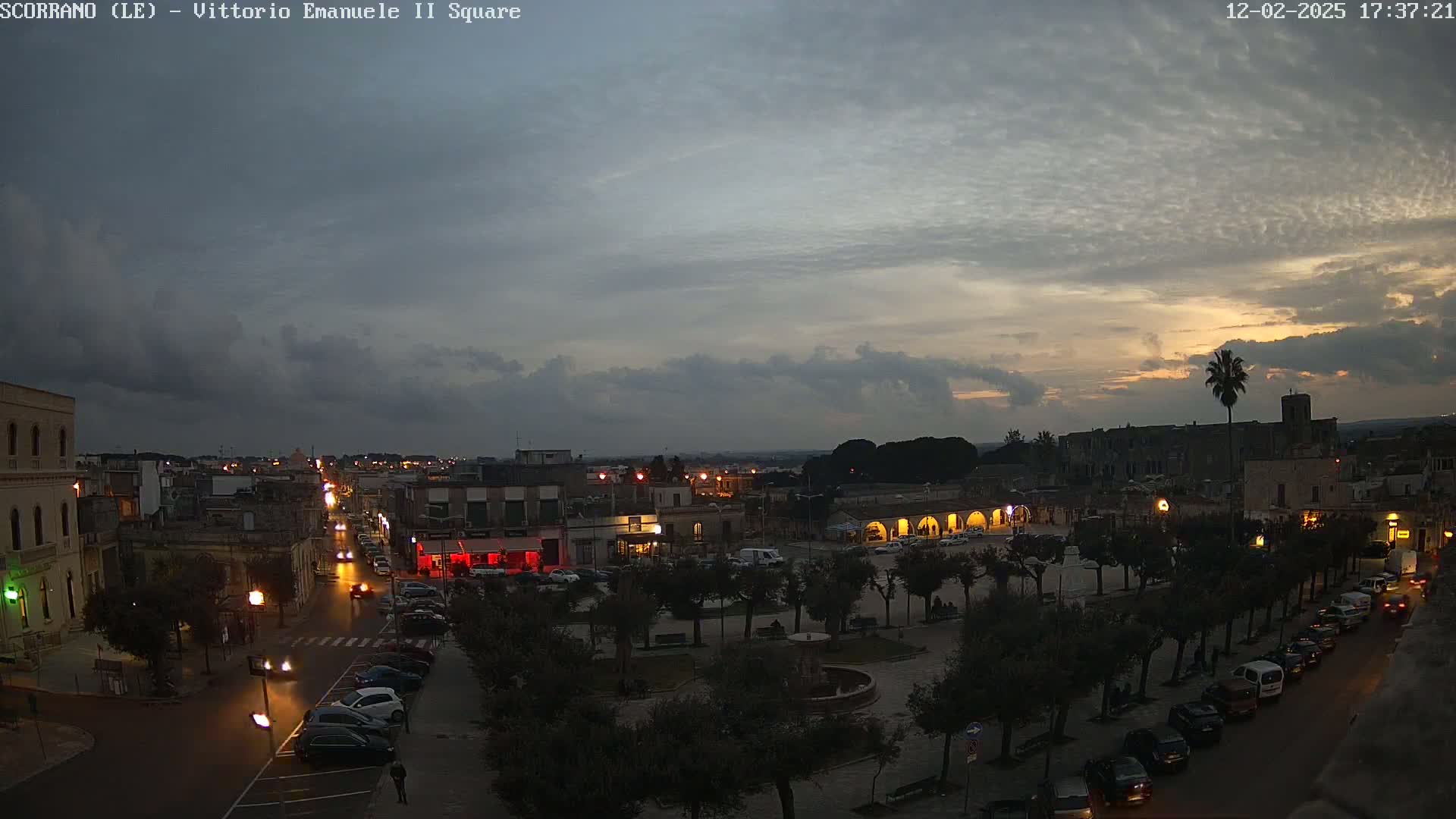 A town square at dusk under a partly cloudy sky, with cars parked along the streets and lights illuminating the buildings and trees.