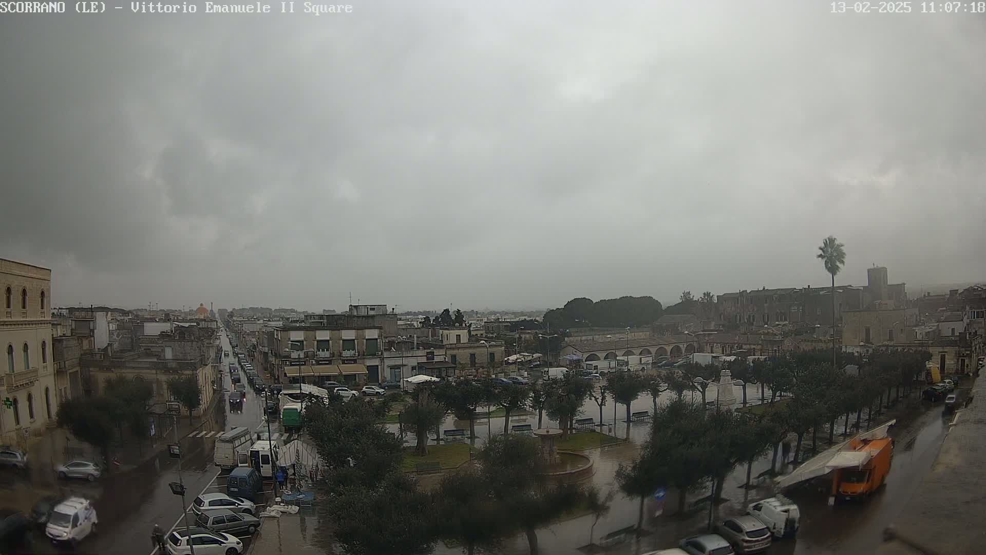 A rain-soaked town square and surrounding streets are viewed from above on an overcast day, showing cars parked and driving through puddles.