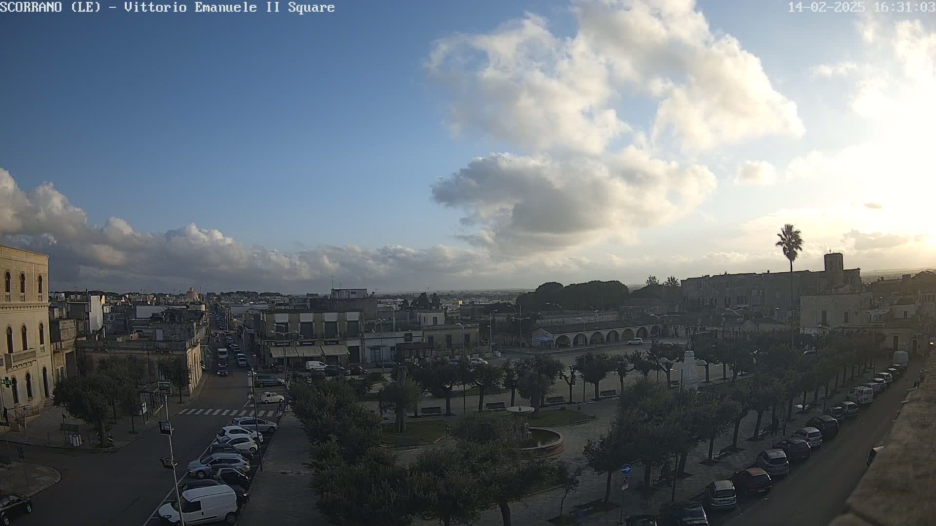 A partly cloudy sky overlooks a town square with parked cars lining the streets and trees in the center.